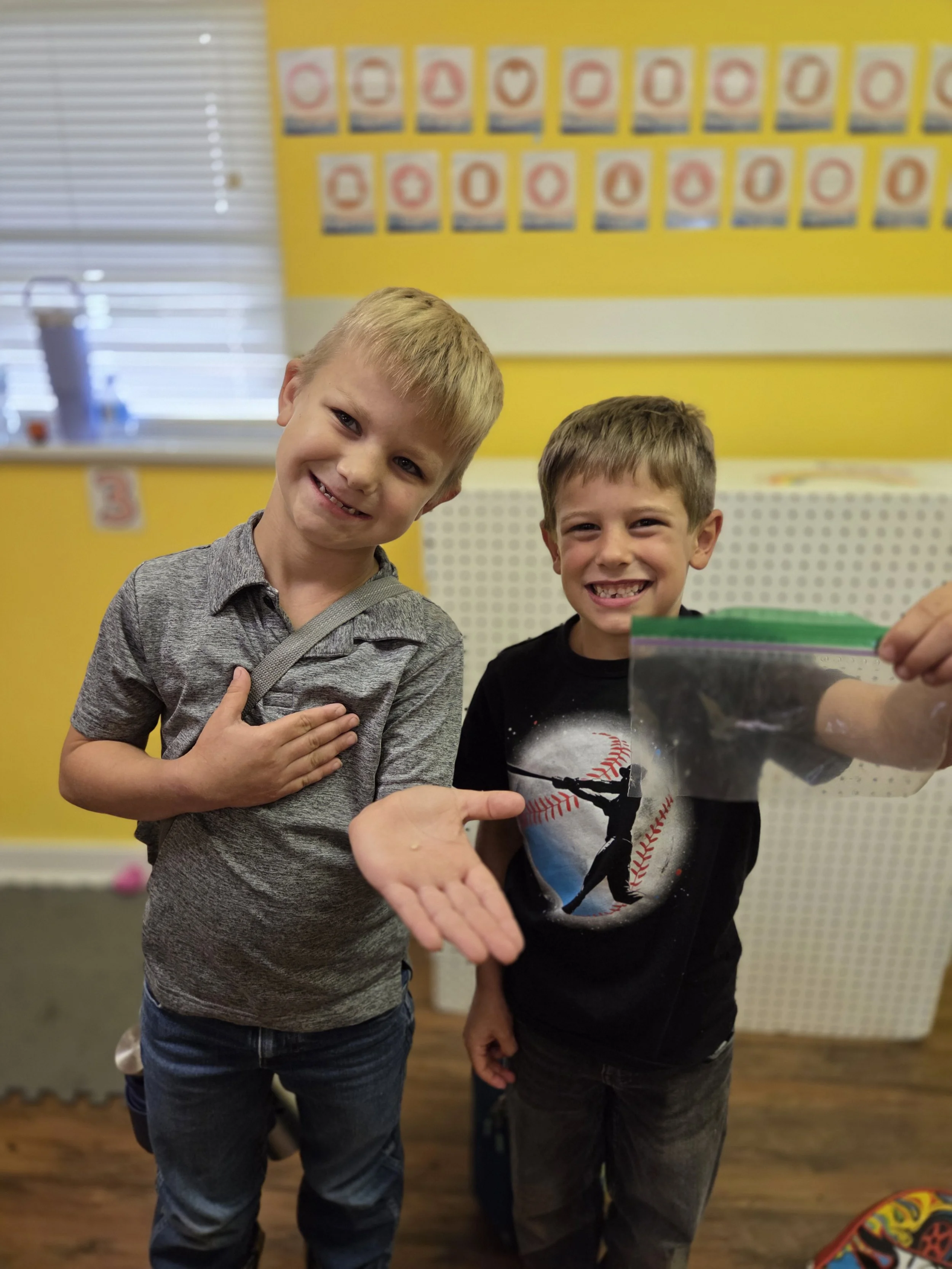 Two young boys smiling and holding up a photograph of another person in a room with educational posters on the yellow wall behind them.