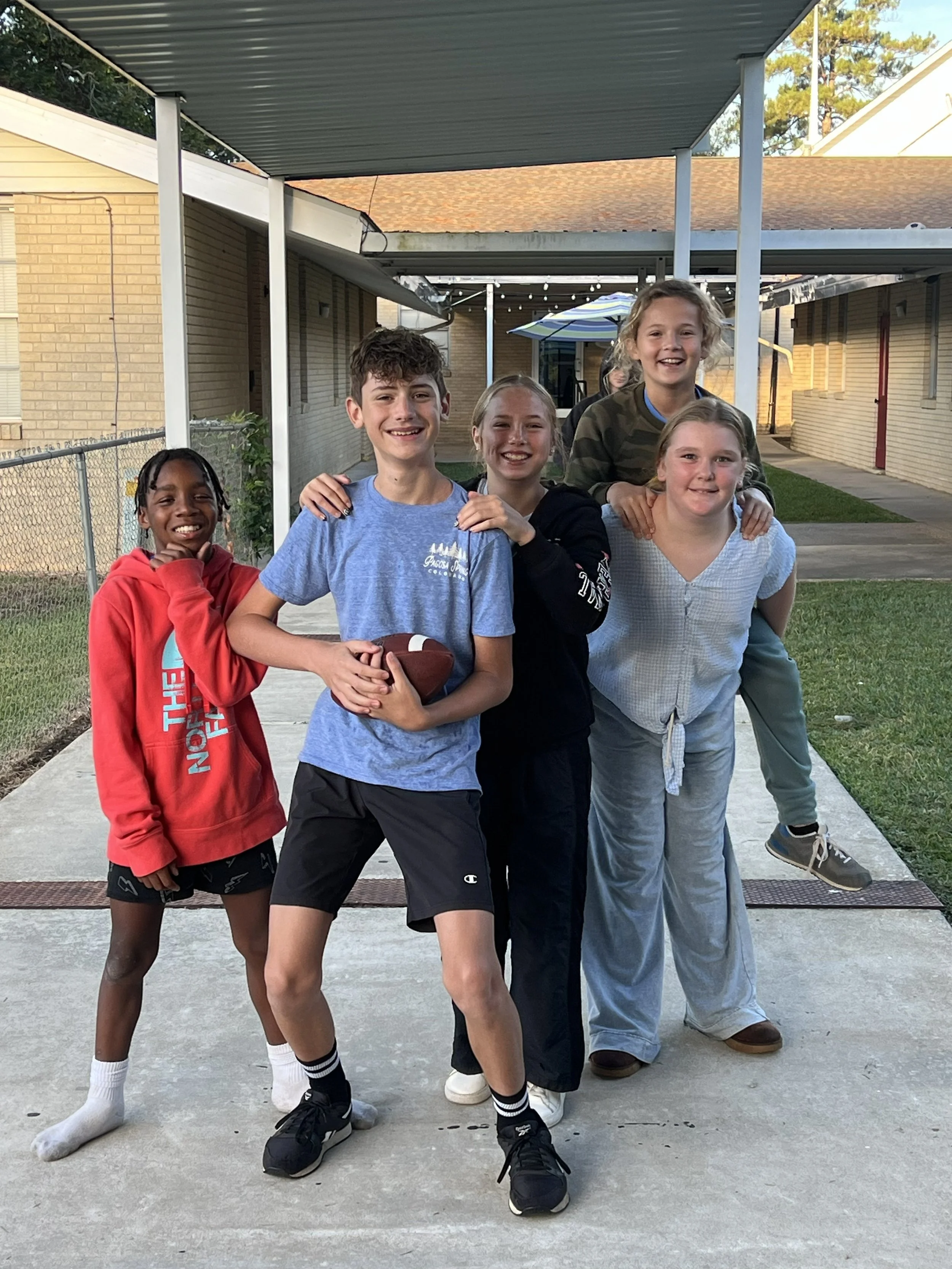 A group of six kids standing outdoors on a concrete sidewalk, smiling and posing together with a fence and buildings in the background, under a covered area with string lights and an umbrella.