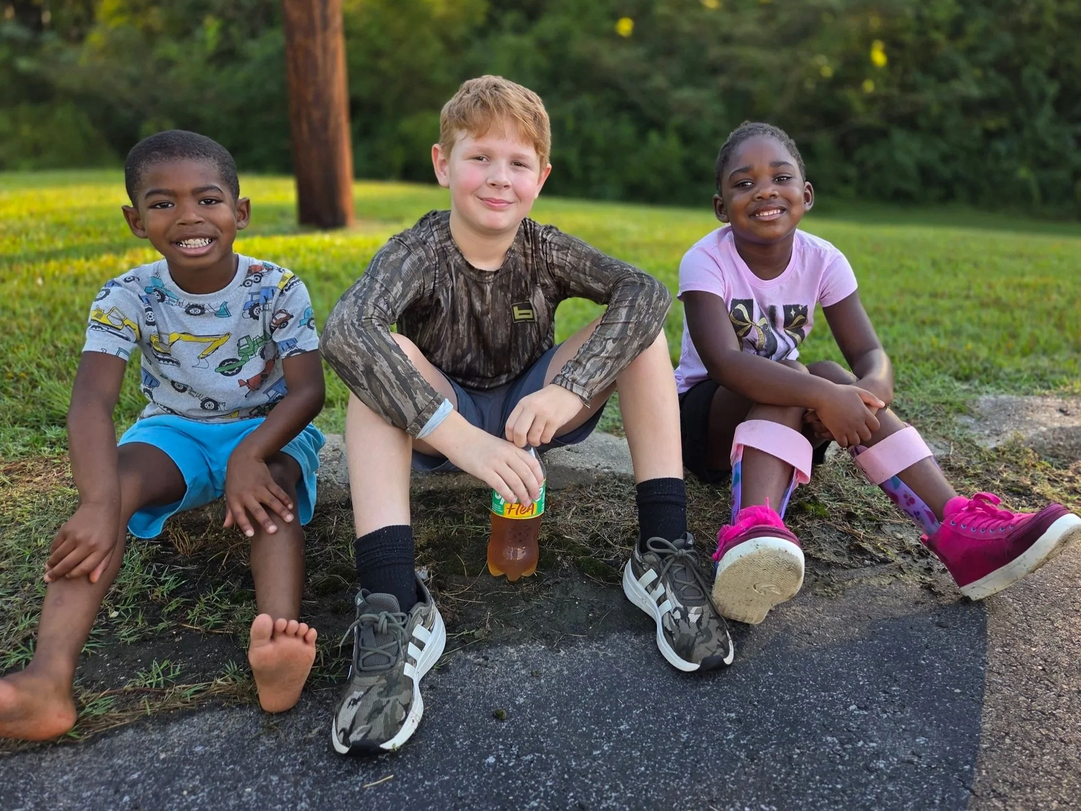 Three children sitting on the ground outdoors in a park with green trees and grass.