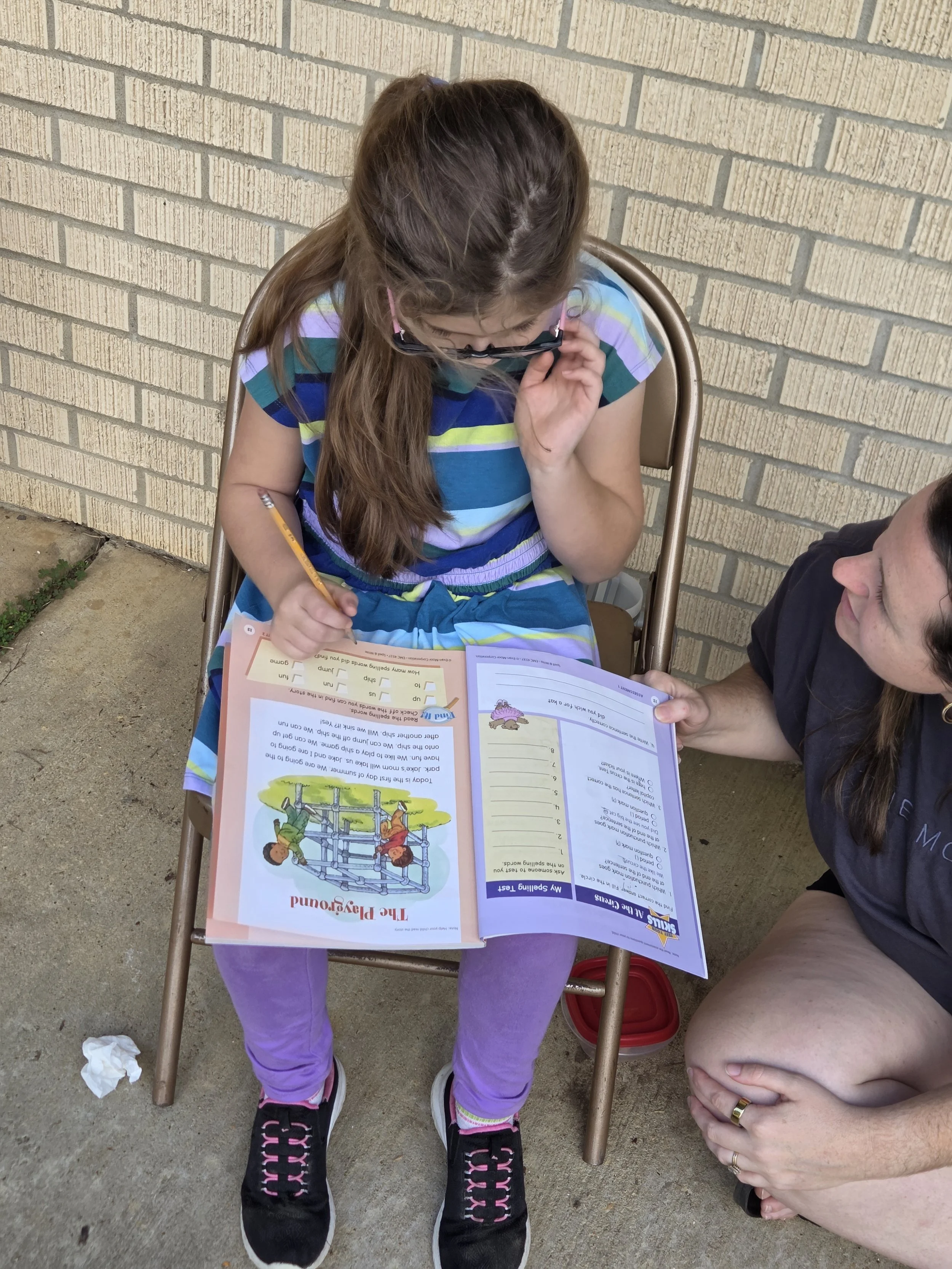 A young girl sitting outdoors on a chair while doing homework with a woman leaning in to help her. The girl is wearing a colorful striped shirt, purple pants, and black sneakers with pink laces. The woman is sitting beside her, holding a pen and helping her with the homework. There is an open workbook on the girl's lap, and they are outside against a brick wall, on a concrete surface.