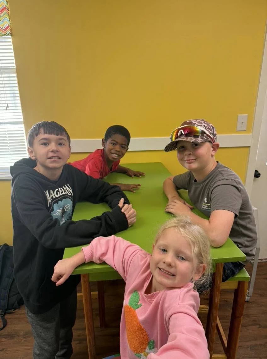 Four children standing around a green table in a room with yellow walls and a window with blinds. One girl in a pink sweatshirt is taking a selfie, smiling at the camera. The other children are boys, with one wearing a camouflage cap and sunglasses, the others in casual clothing.