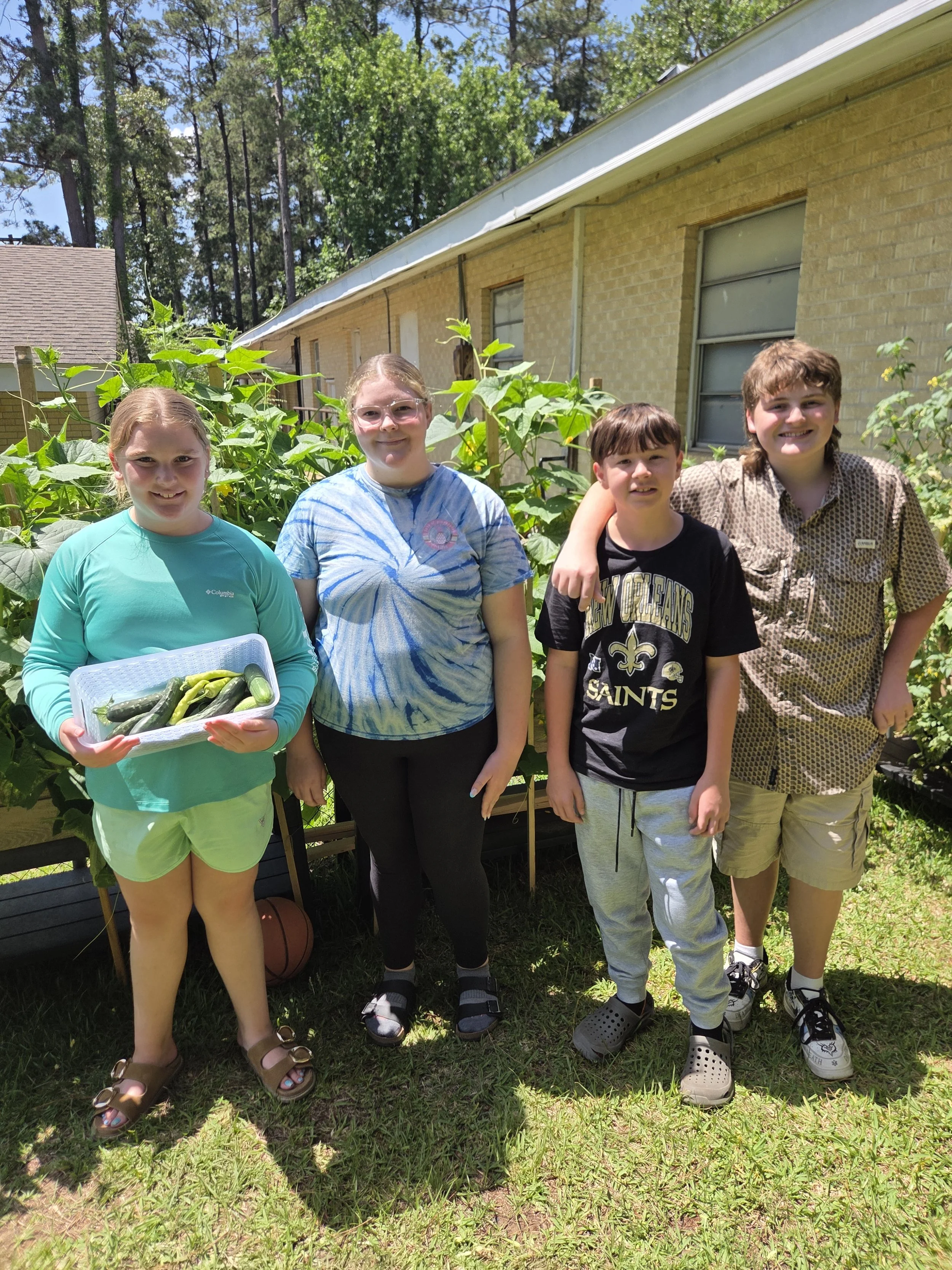 Four children stand outdoors in front of a garden with green leafy plants, smiling at the camera. The girl on the left holds a plastic tray of cucumbers. The background includes a yellow brick house, trees, and a bright blue sky.