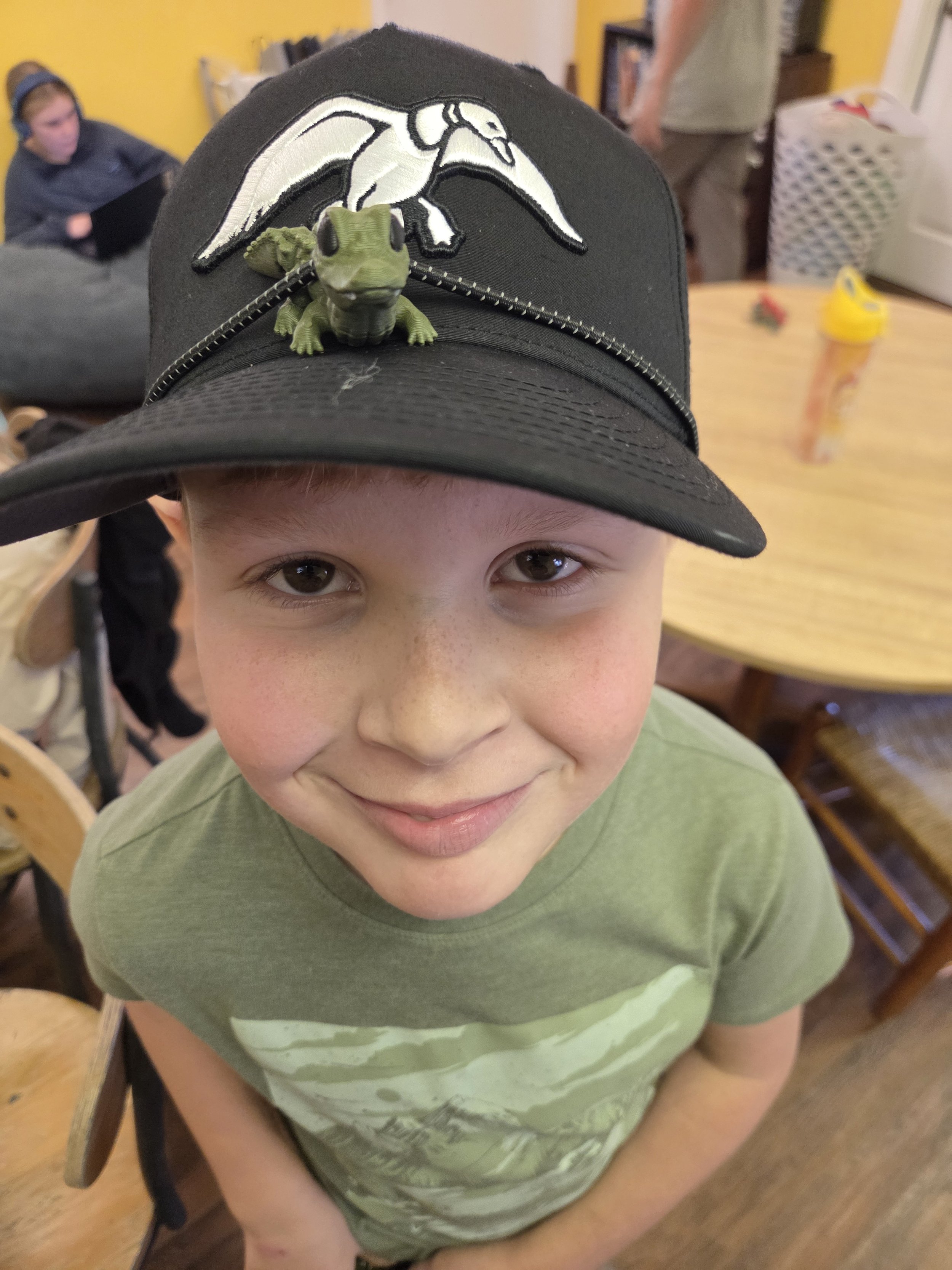 A boy wearing a black cap with a white heron and a small green toy dinosaur on top of it, smiling inside a room with a yellow wall and a wooden table.