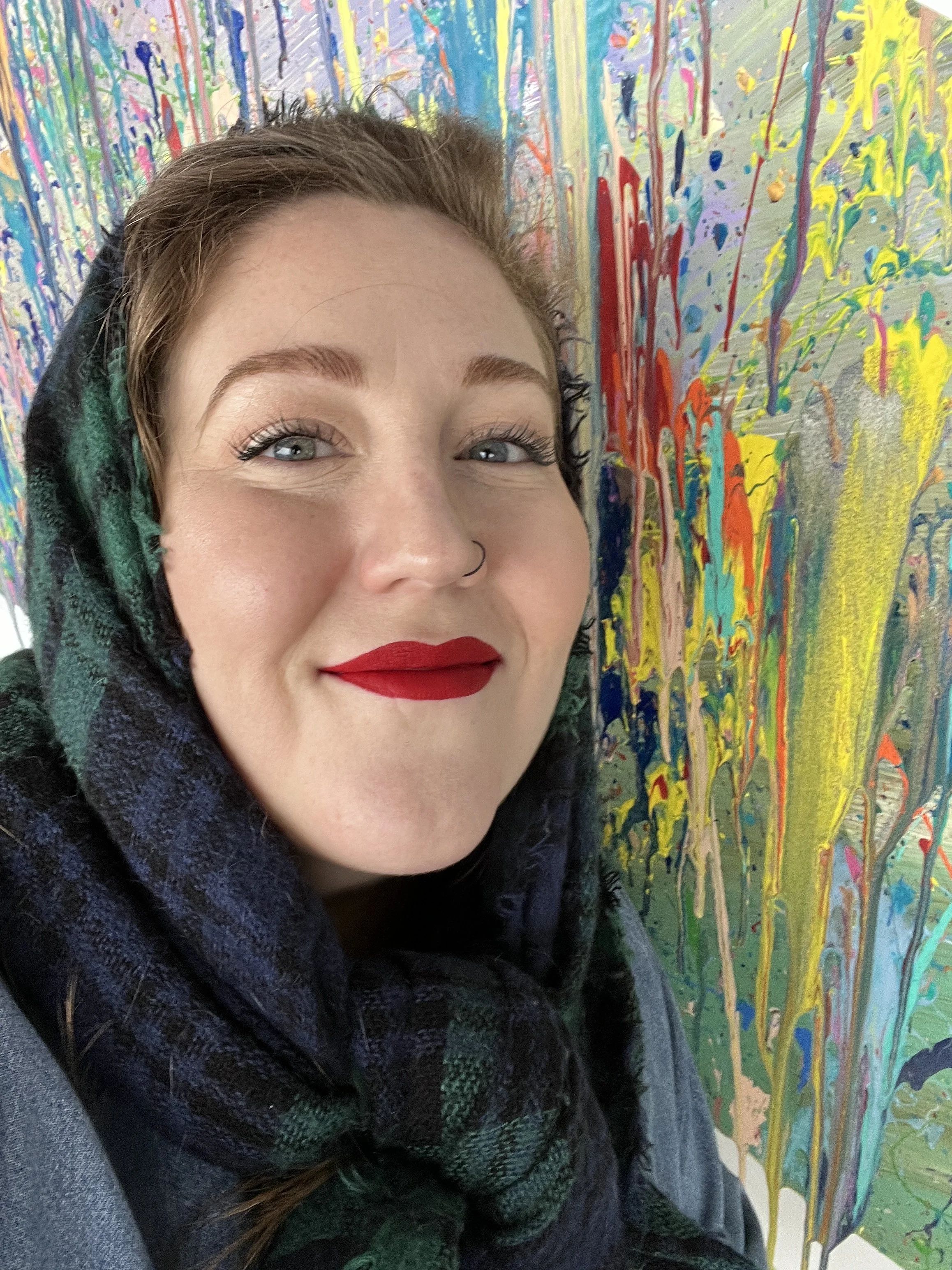 Close-up of a smiling woman with red lipstick, light eye makeup, and a nose ring, wearing a dark hoodie and scarf, standing in front of an abstract colorful painting with splashes of paint in various colors.