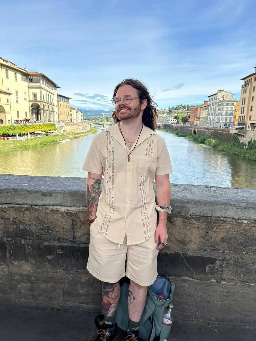 A man with long wavy hair and glasses stands smiling in front of a river with historic buildings on both sides. He is wearing a beige short-sleeved shirt and shorts, with tattoos visible on his arms and legs. A backpack and a water bottle are on the ground beside him.