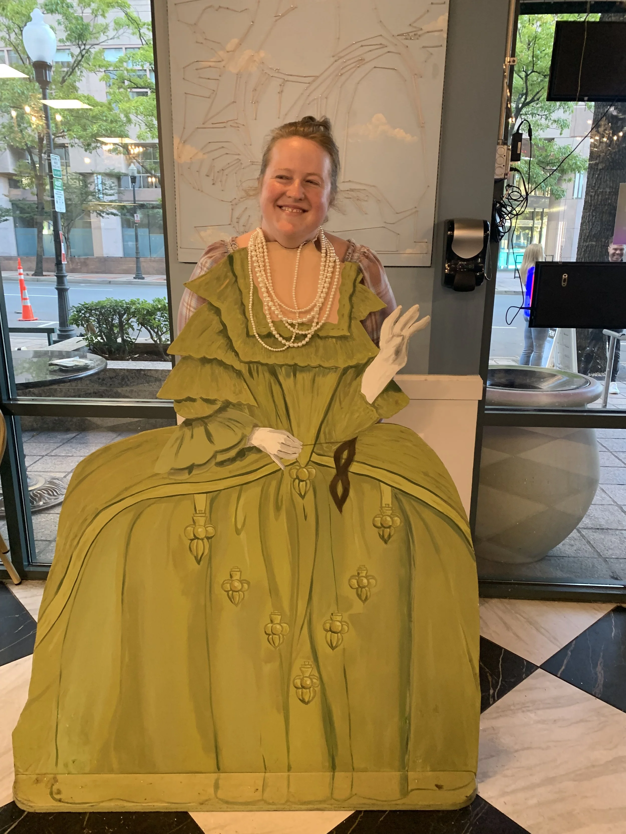 A woman smiling through a cutout of a yellow dress. The cutout is part of a display in a store with a window showing a street scene outside.