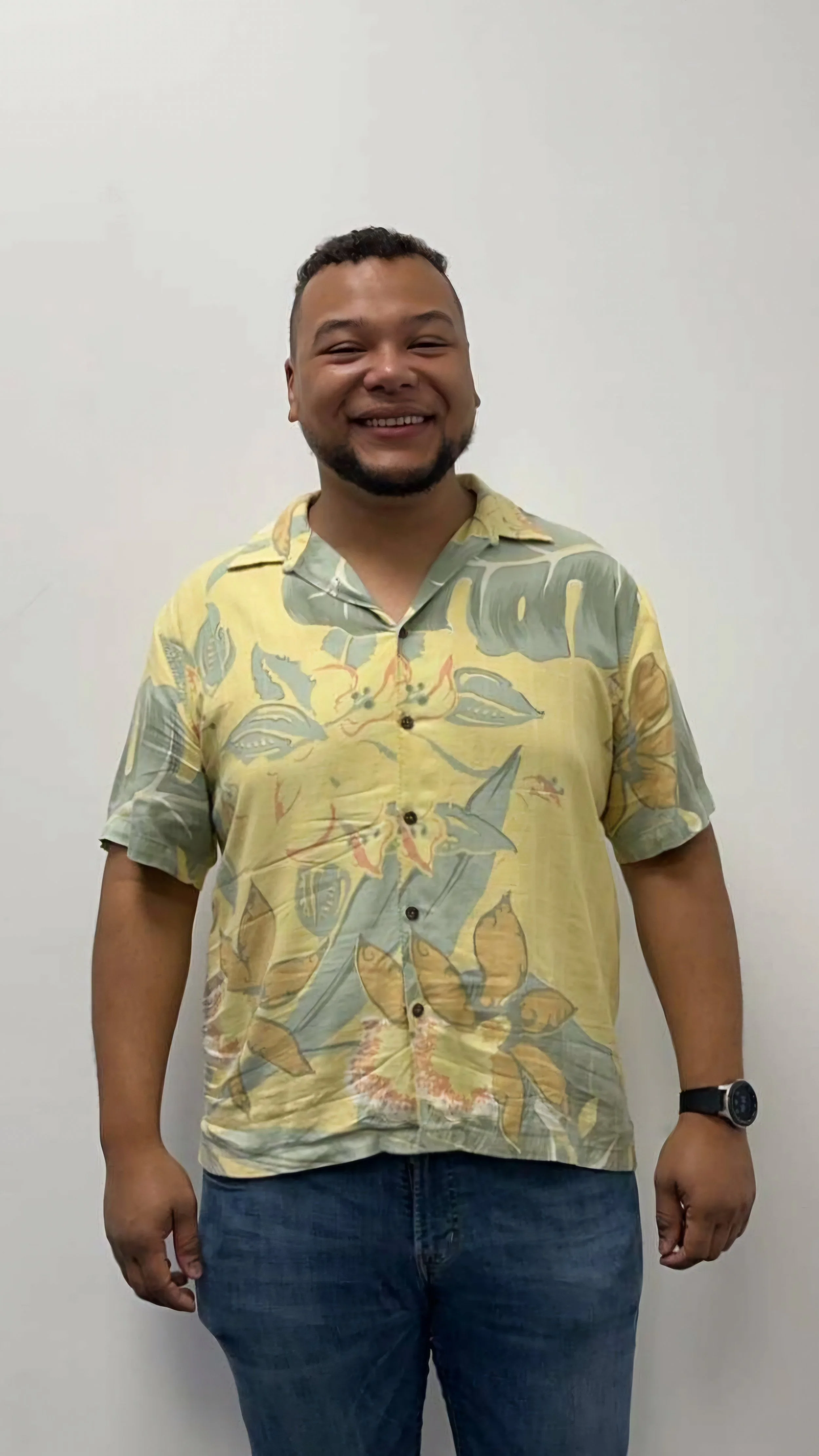 A smiling man wearing a yellow Hawaiian shirt with floral and leaf patterns, standing against a plain white wall.