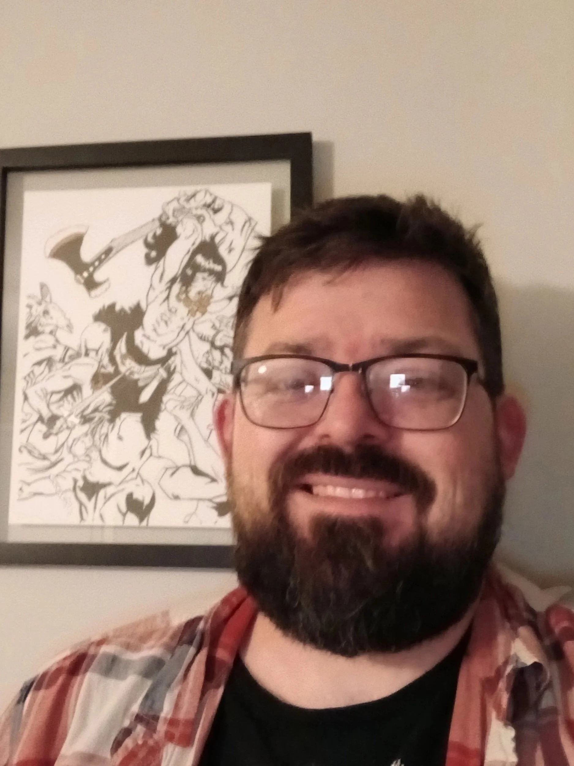 A man with glasses and a beard smiling, wearing a plaid shirt over a black t-shirt, standing in front of a framed black and white illustration of a mythical creature on a beige wall.
