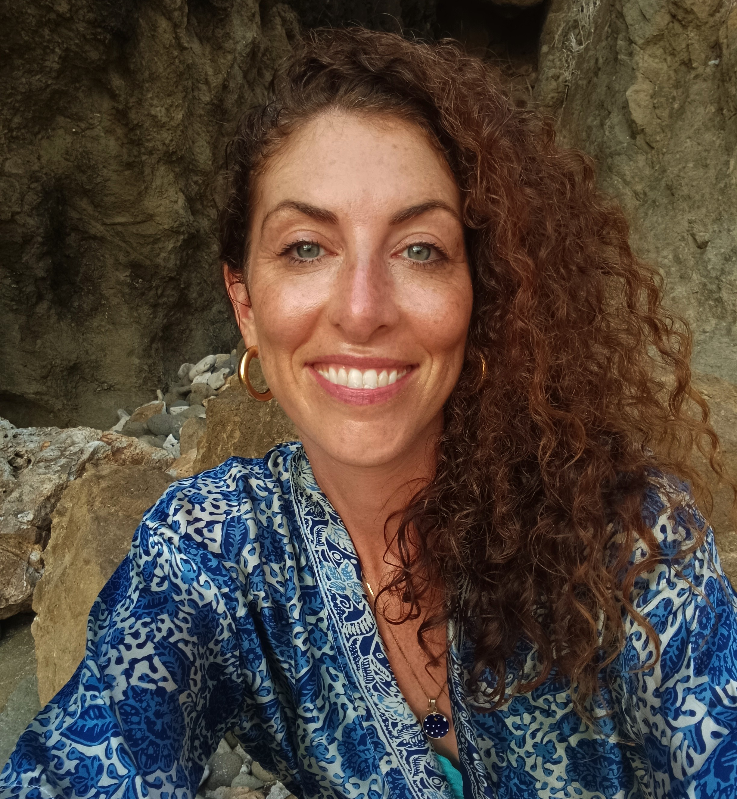 Close-up of a smiling woman with curly red hair and blue eyes, wearing gold hoop earrings, a gold necklace with a pendant, and a blue and white patterned shirt. She is outdoors against a rocky background.