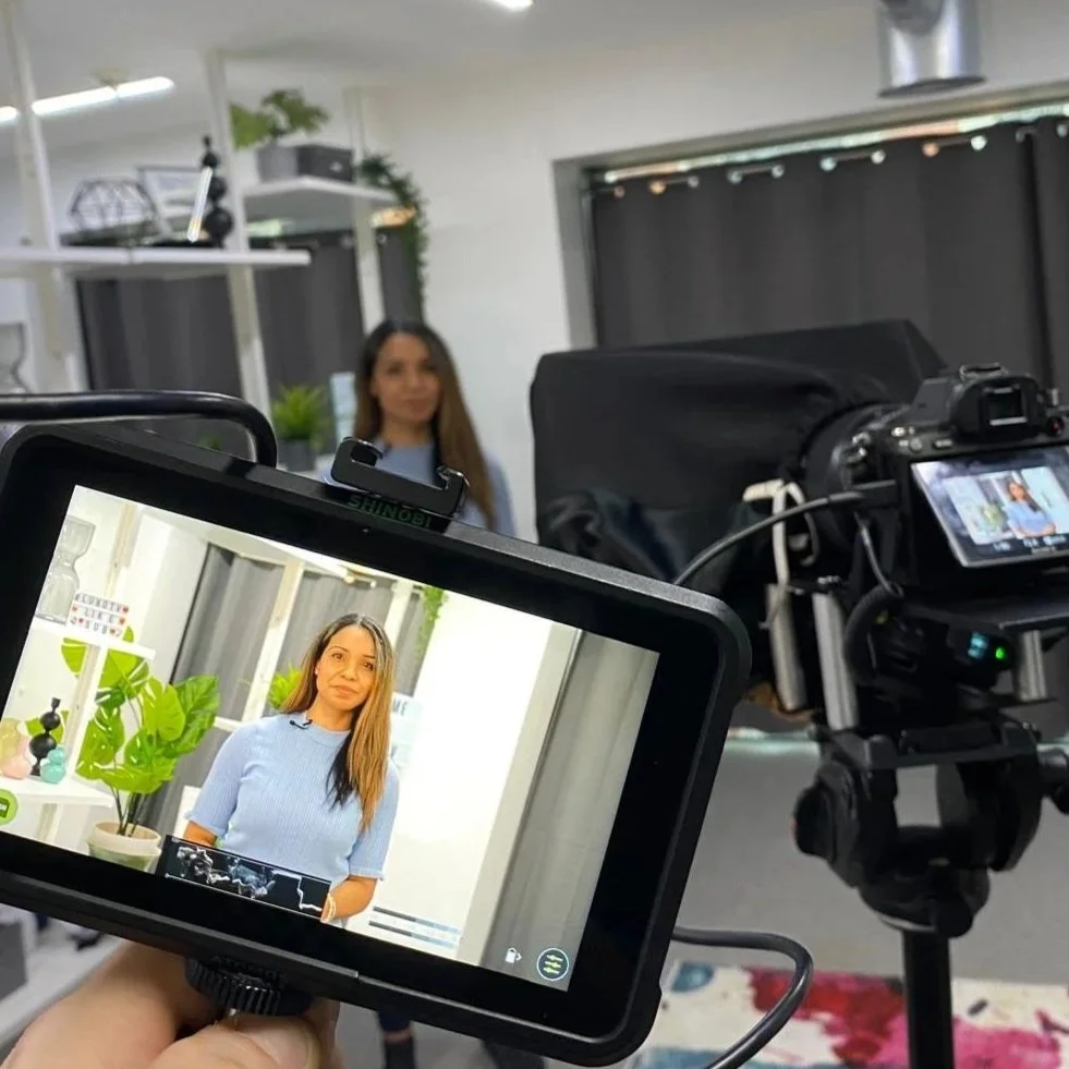 A woman is being filmed in a studio with a backdrop and decorative plants, and her image is displayed on a camera monitor.