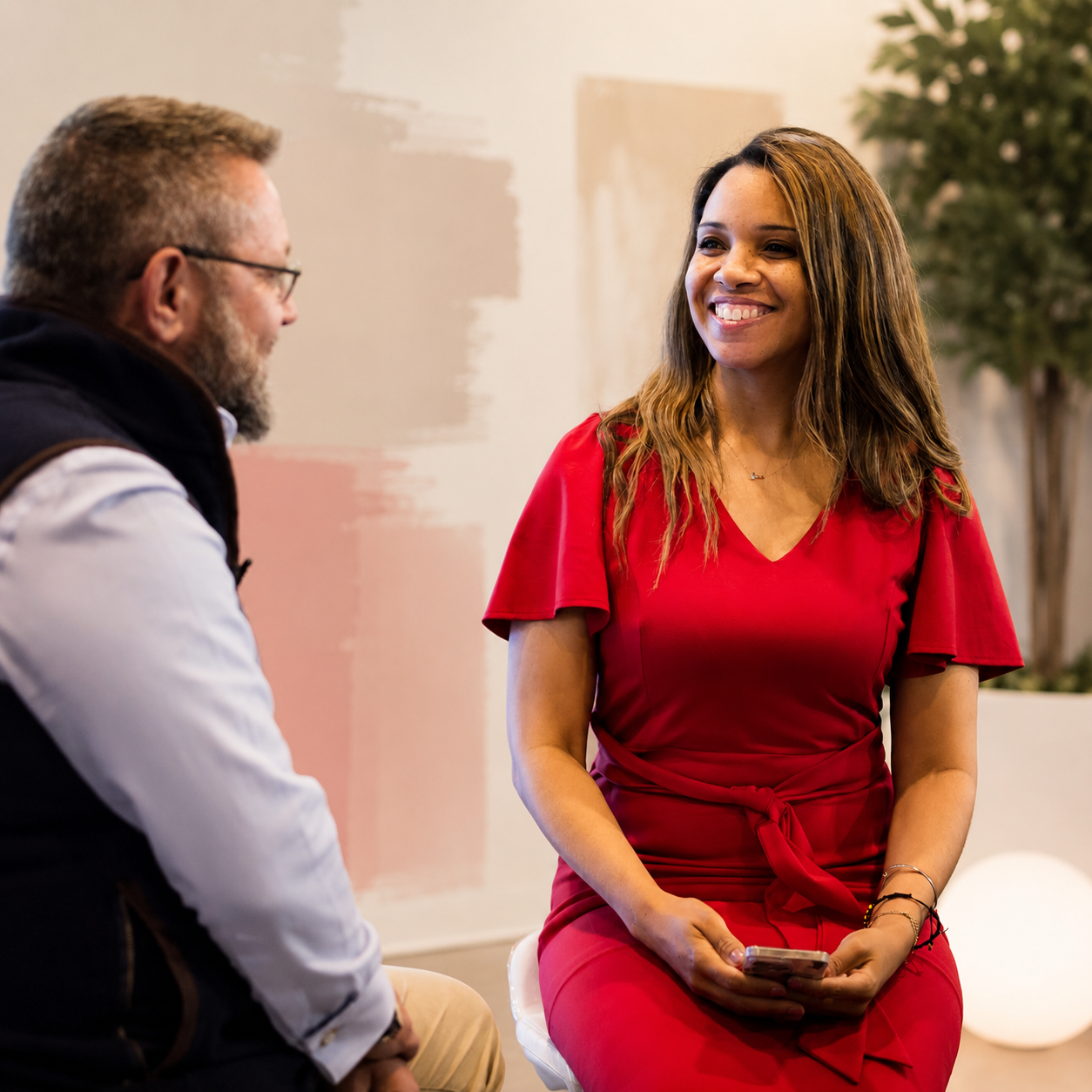 A woman in a red dress sitting and holding a smartphone, smiling as she talks to a bearded man wearing glasses and a black vest.