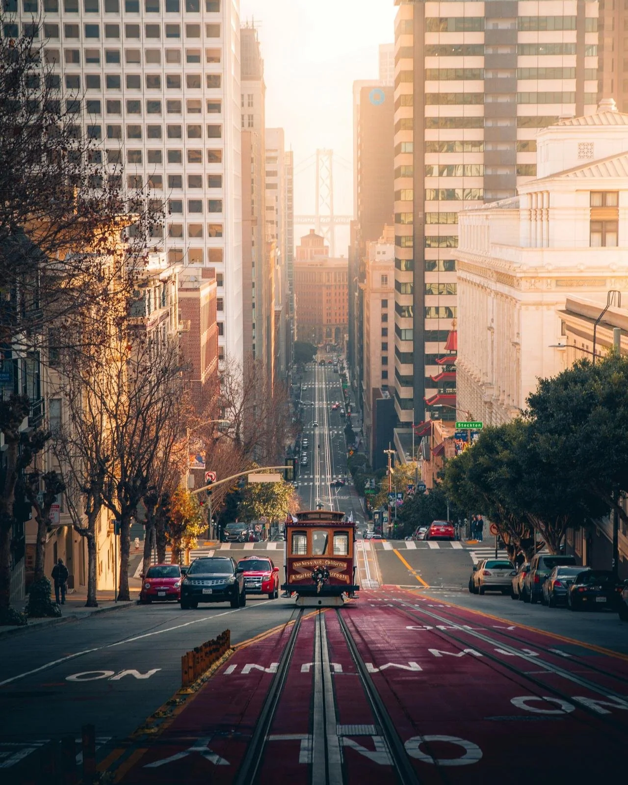 A San Francisco cable car on a hilly city street surrounded by tall buildings with a view of the Bay Bridge in the distance.