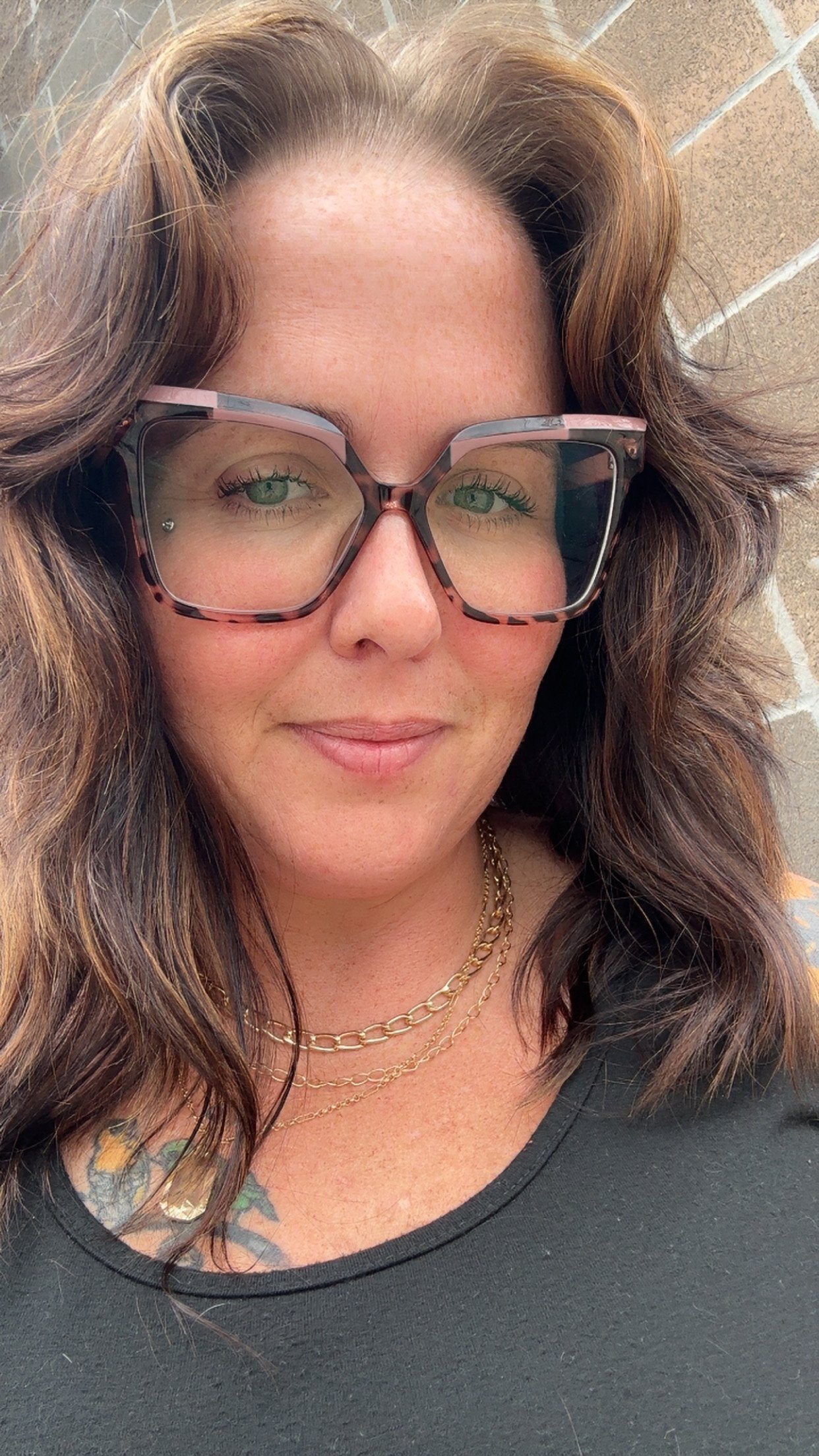 Close-up of a woman with wavy brown hair, wearing large sunglasses with a pink and tortoise shell frame, gold layered necklaces, and a dark top, standing against a brick wall.