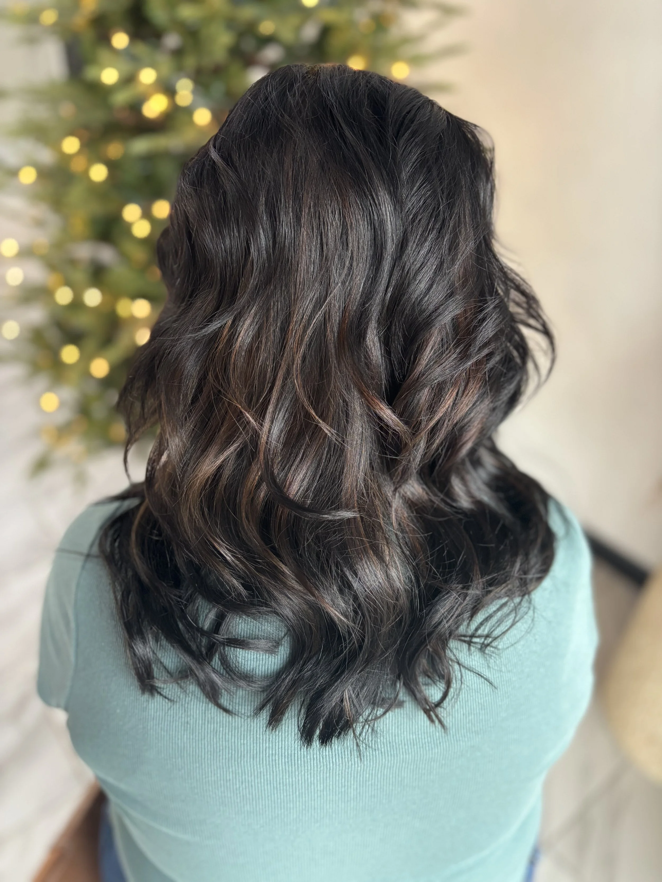 Back view of a woman with wavy, shoulder-length black hair wearing a light blue top, with a background featuring blurred holiday tree lights.