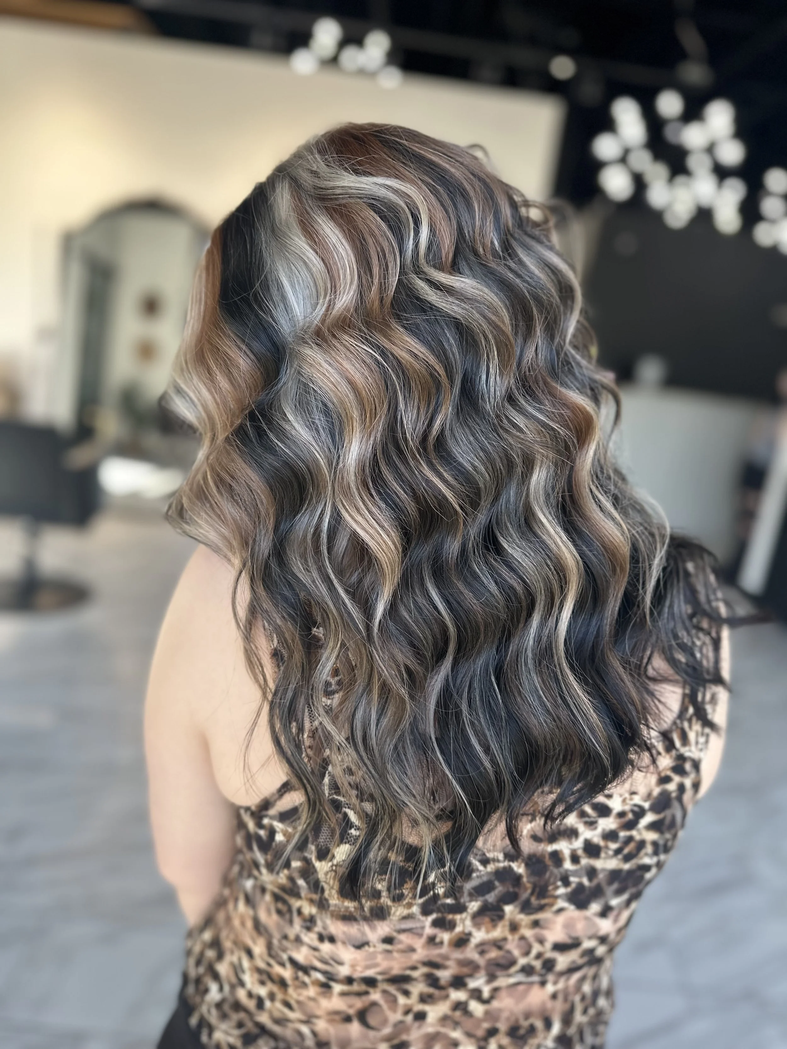 Woman with long, wavy hair featuring highlights in shades of blonde, brown, and black, wearing a leopard print top, in a salon setting.