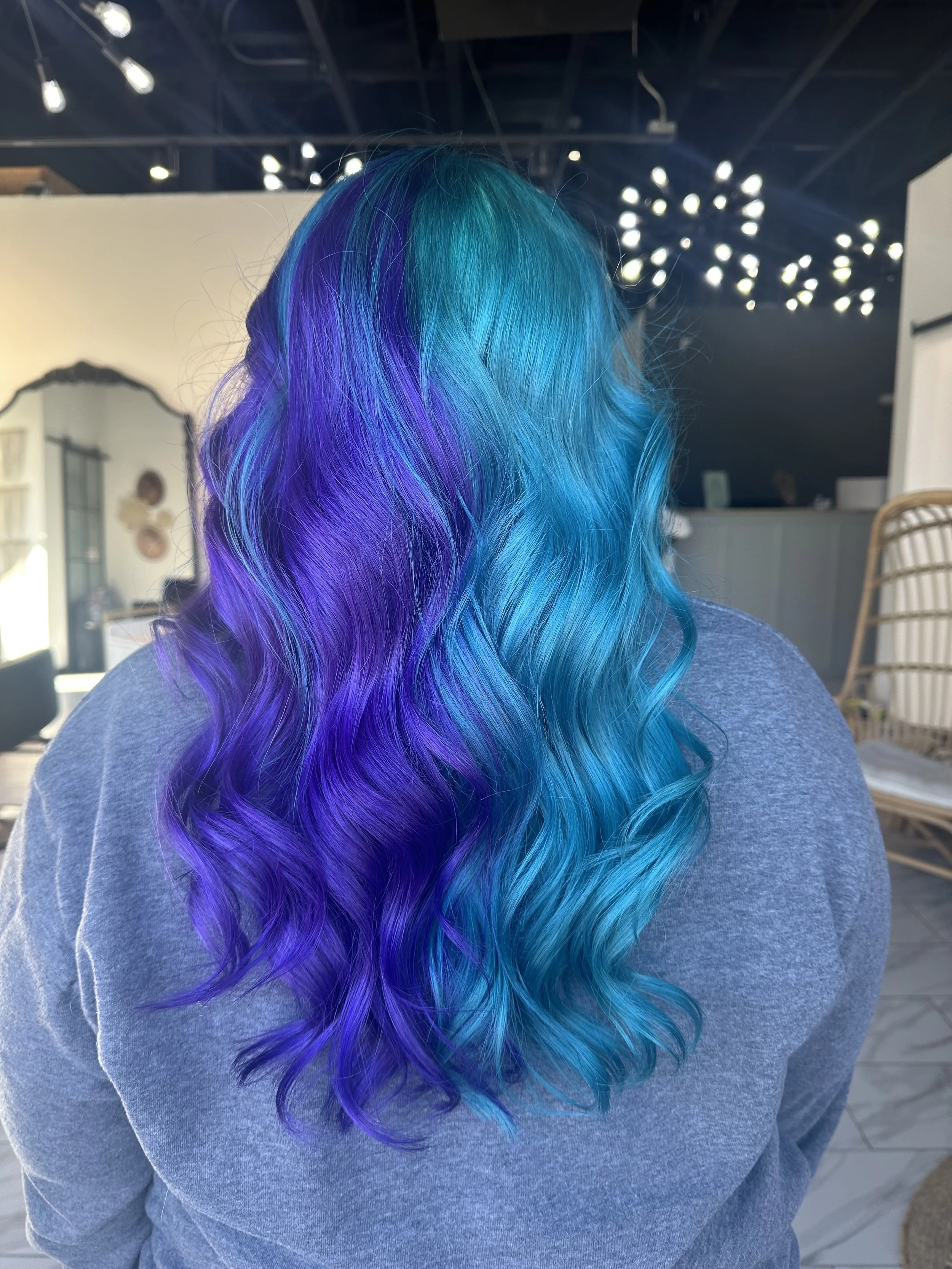 Back view of a person with long, wavy hair dyed in vibrant shades of purple, blue, and teal, wearing a gray shirt, in a salon setting.