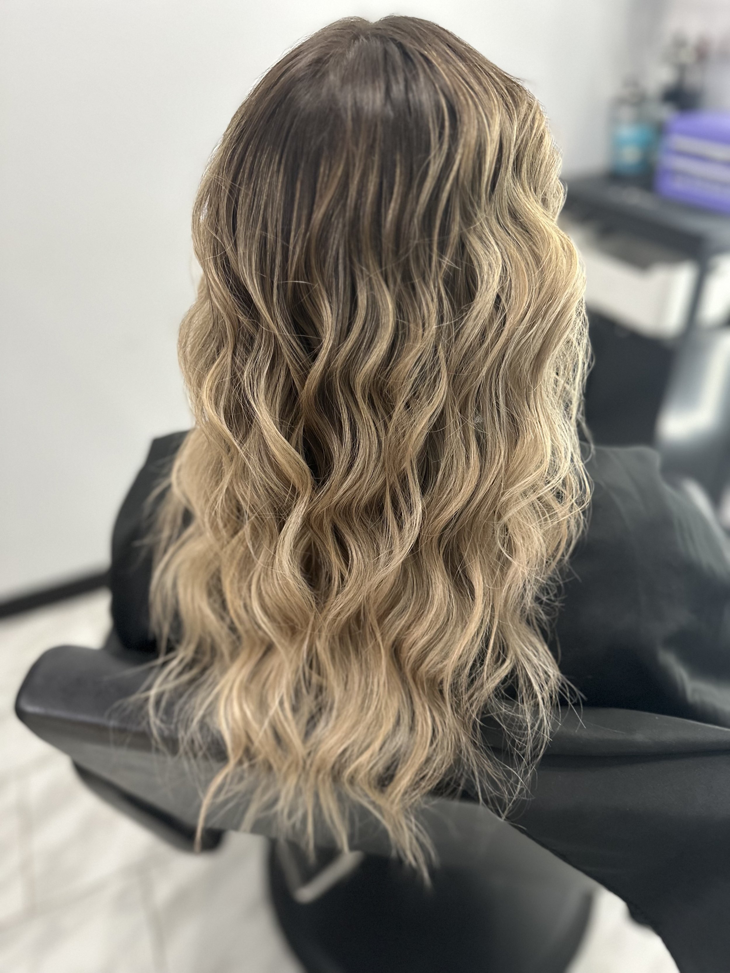 Back view of a person with long, wavy blonde hair sitting in a salon chair.