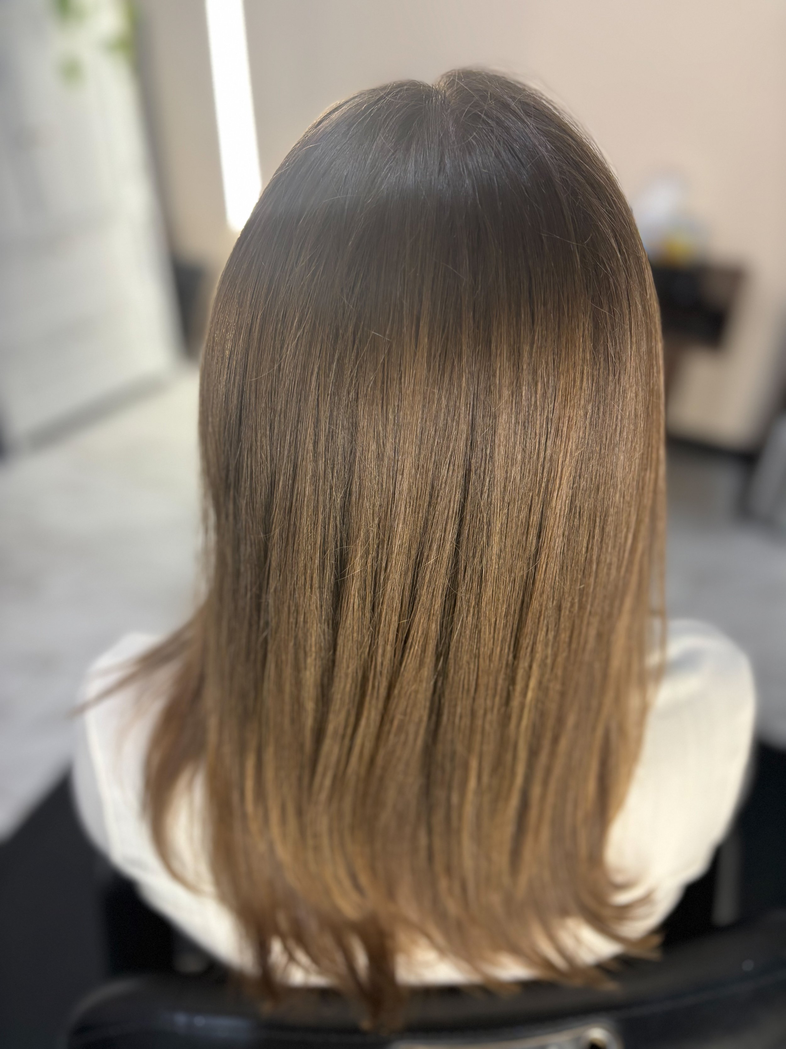 Back view of a person's head with long, straight, light brown hair, with darker roots, sitting in a salon chair.
