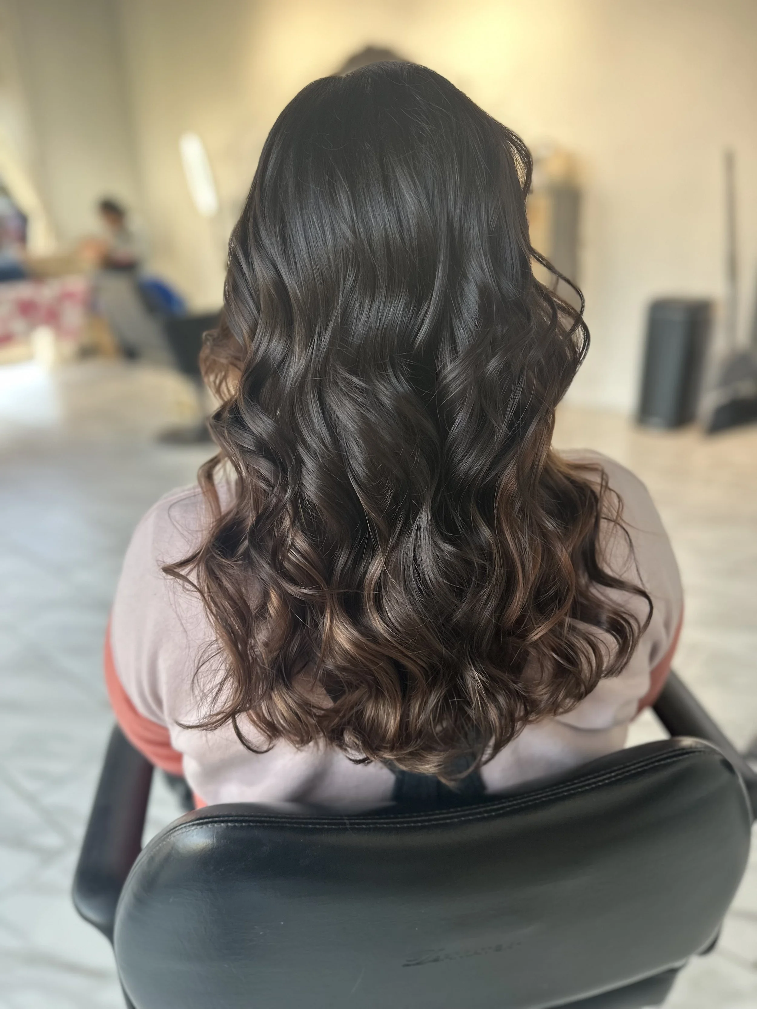 Back of a woman with long, dark, wavy hair sitting in a salon chair.