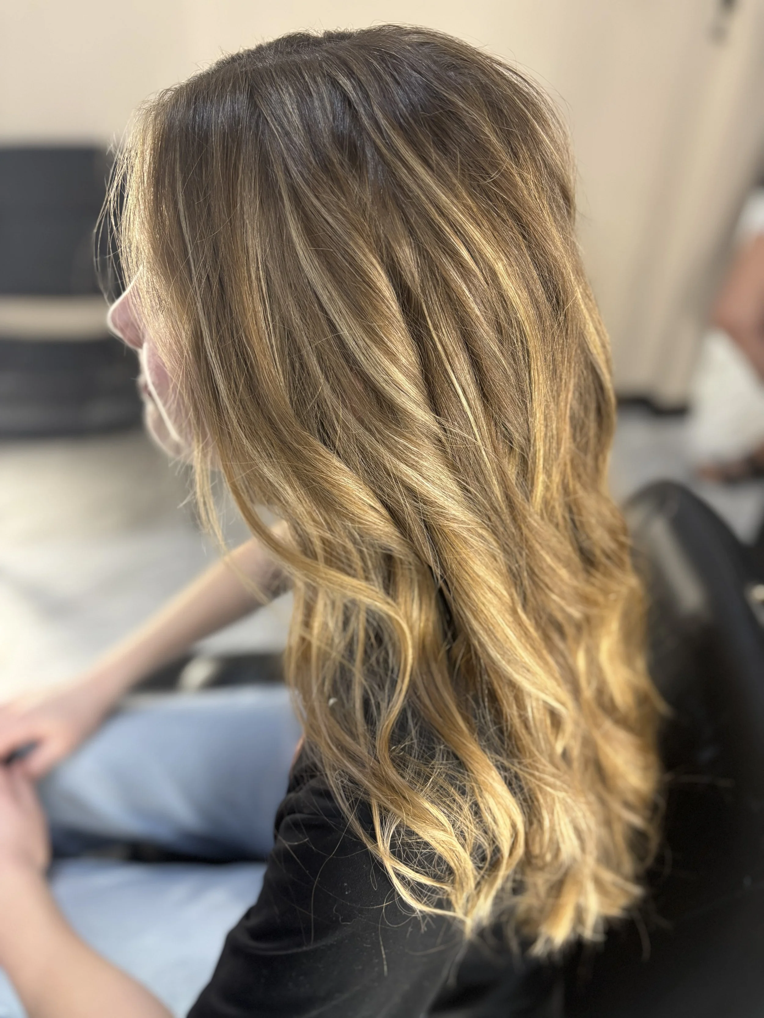 A person with wavy, blonde hair with darker roots, seen from the side, sitting in a room with beige walls and wearing a black top.