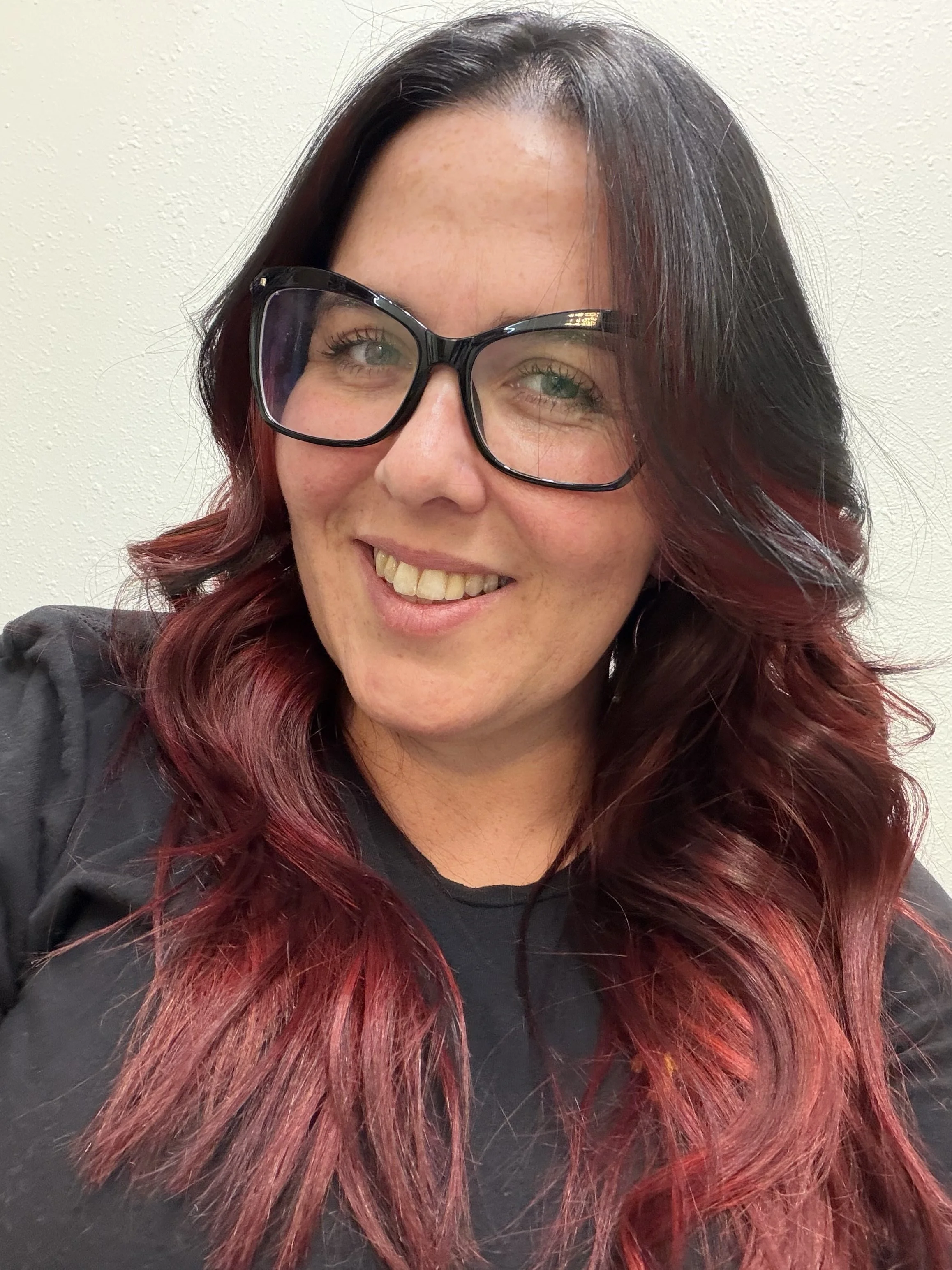 A woman with long, wavy hair that is black at the top and transitions to red at the ends, wearing large black glasses, smiling against a plain, light-colored wall.