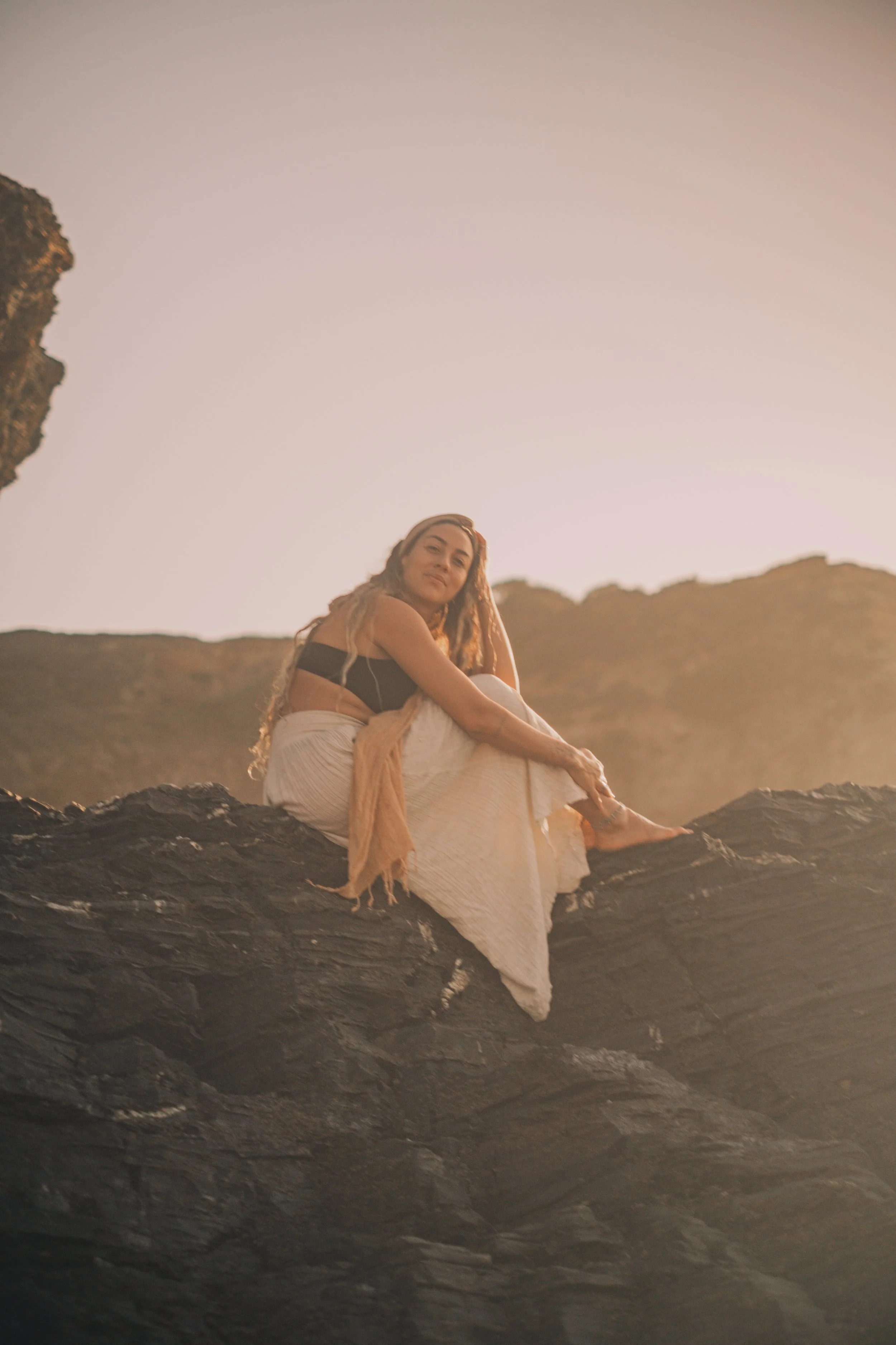 A woman sitting on a rock formation outdoors at sunset, wearing a black top and a long white skirt, with a light brown scarf, relaxed and smiling.
