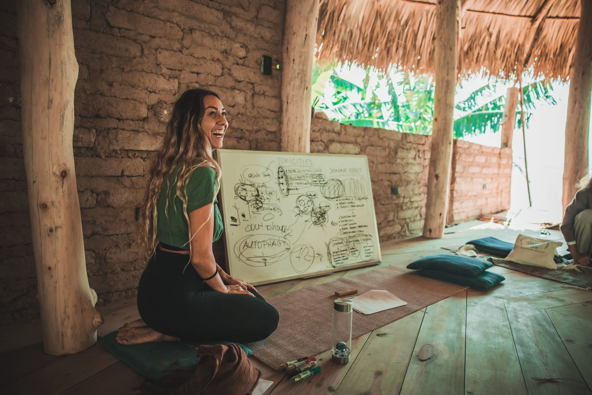 A woman with long wavy hair, wearing a green top, kneeling on a cushion and smiling in front of a whiteboard with handwritten notes and diagrams, inside a rustic open-air structure with bamboo and wooden beams, tropical plants visible outside.