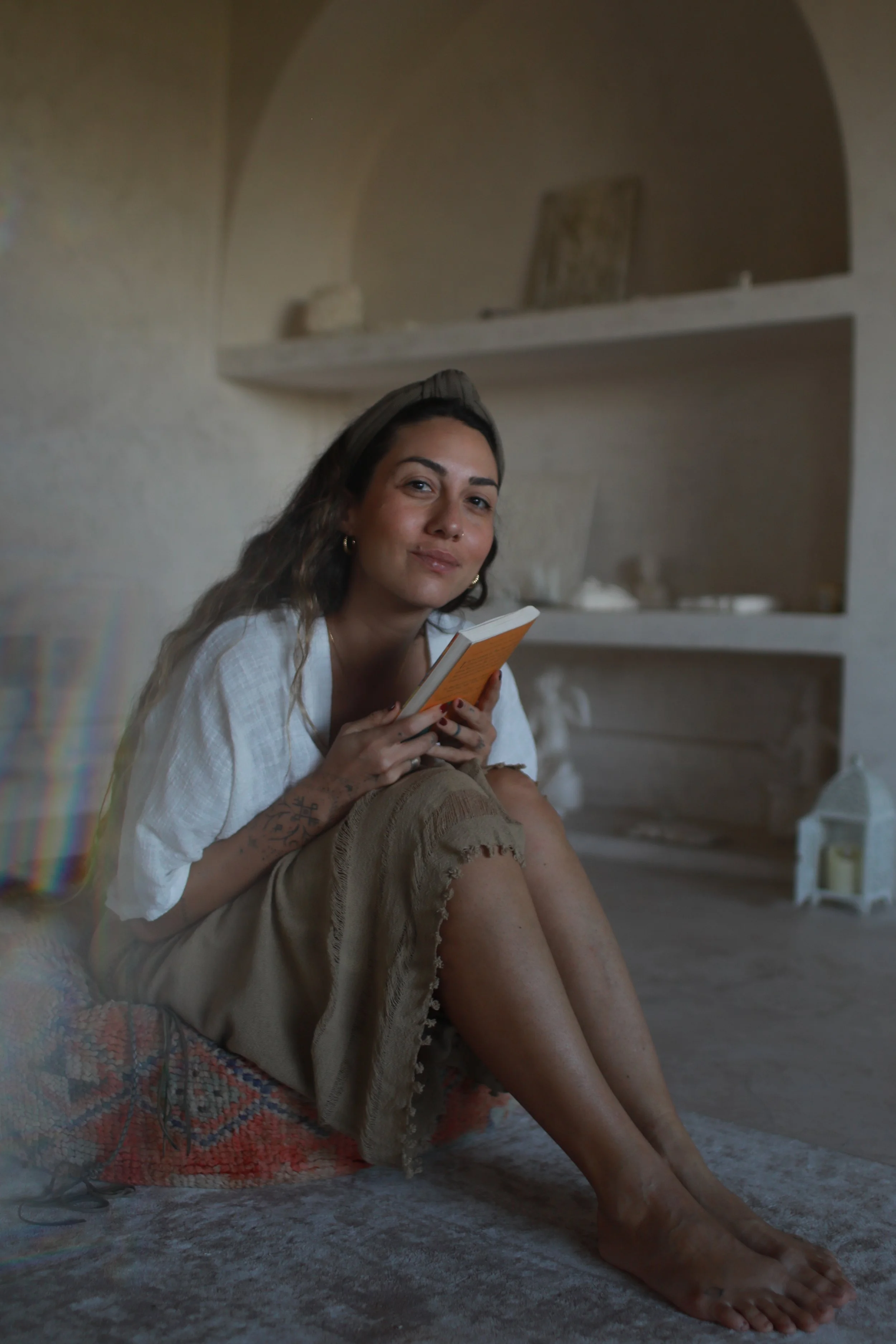 A woman sitting on a blanket, reading a book, in a room with minimalist decor and built-in shelves.