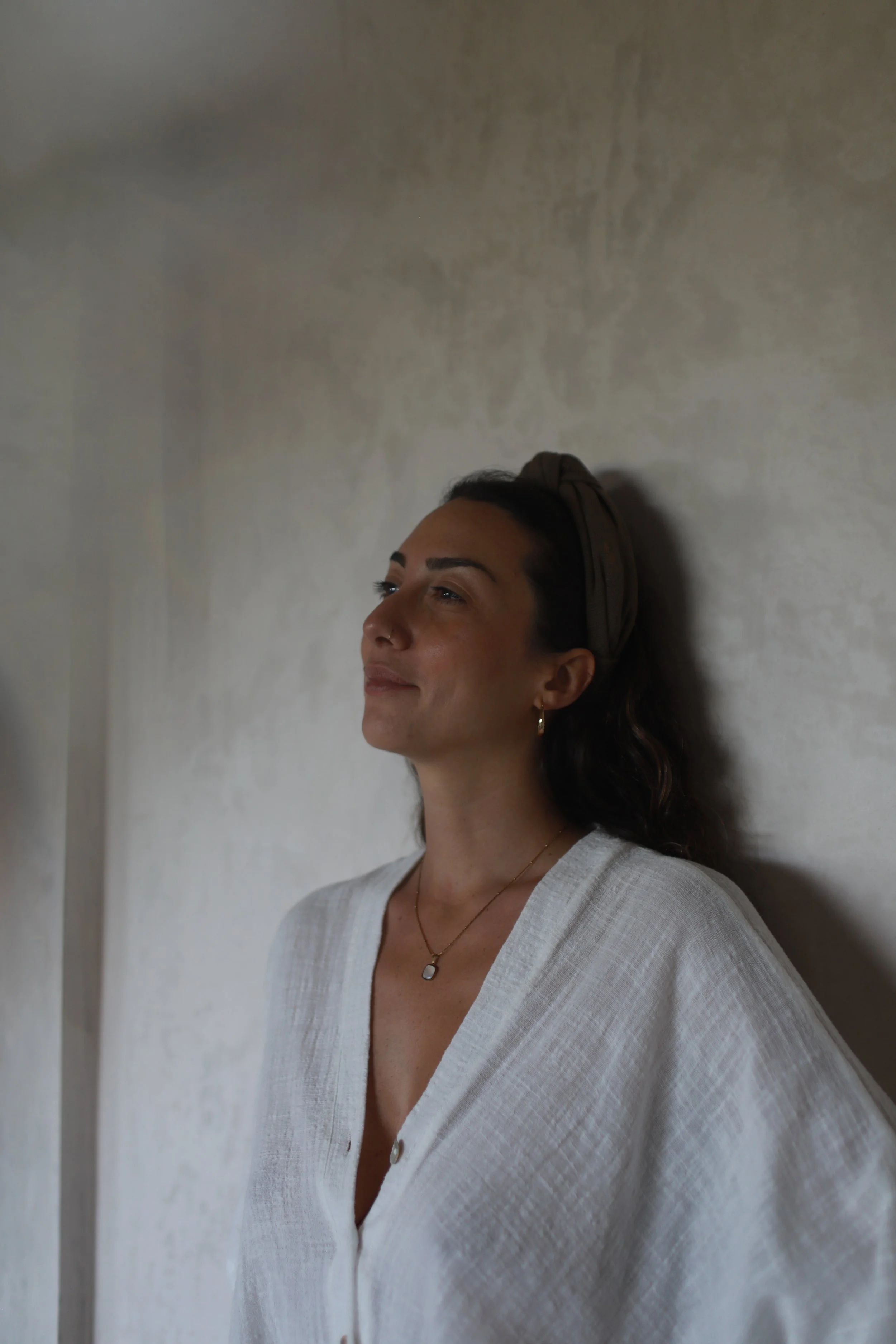 A woman with dark hair, a headband, earrings, and a nose piercing is leaning against a textured white wall, with her eyes closed and a slight smile on her face.