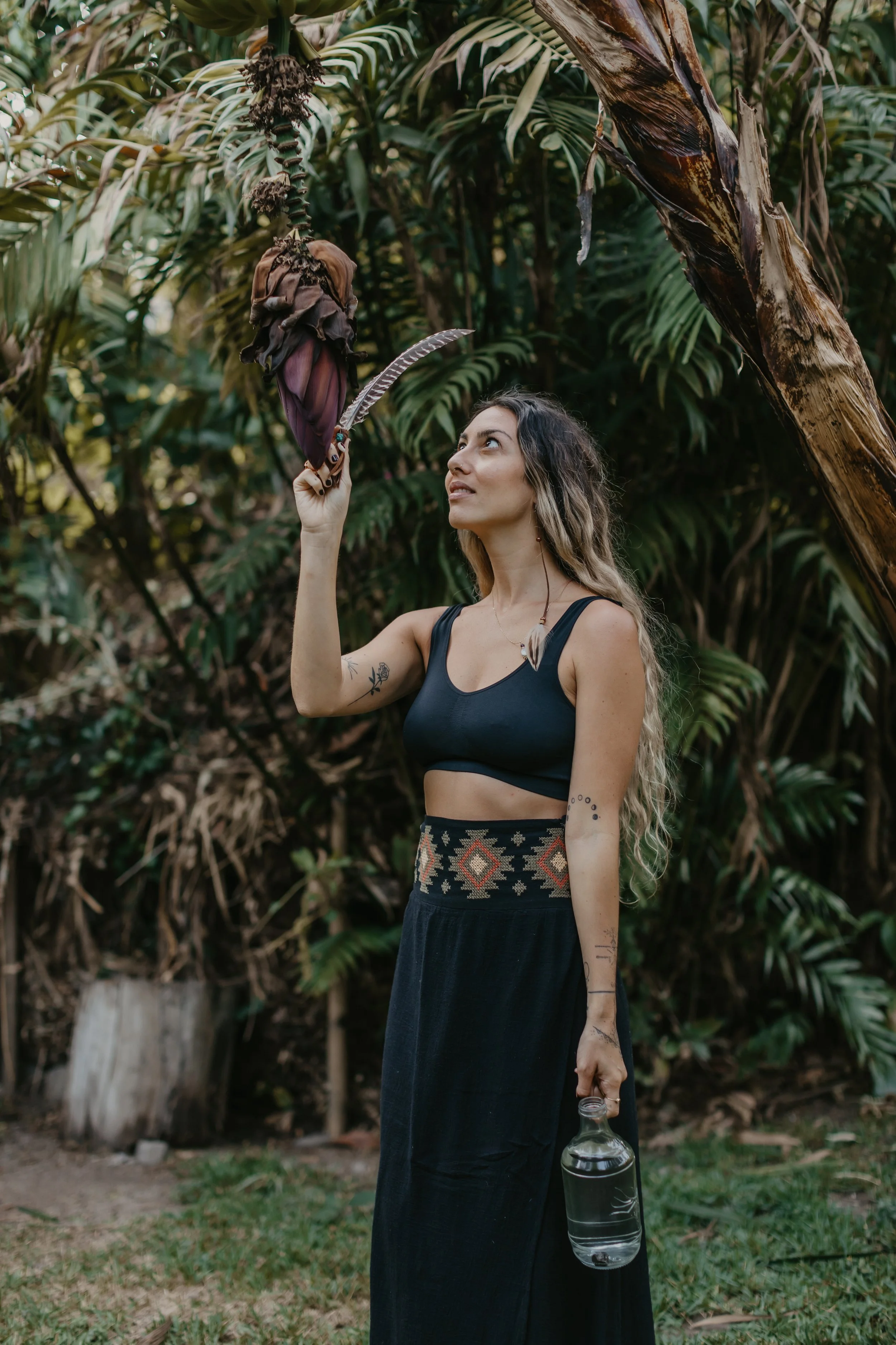 A woman with long wavy hair and tattoos on her arms holds a large moth on her finger in a lush, tropical setting with tall green plants and a banana tree.
