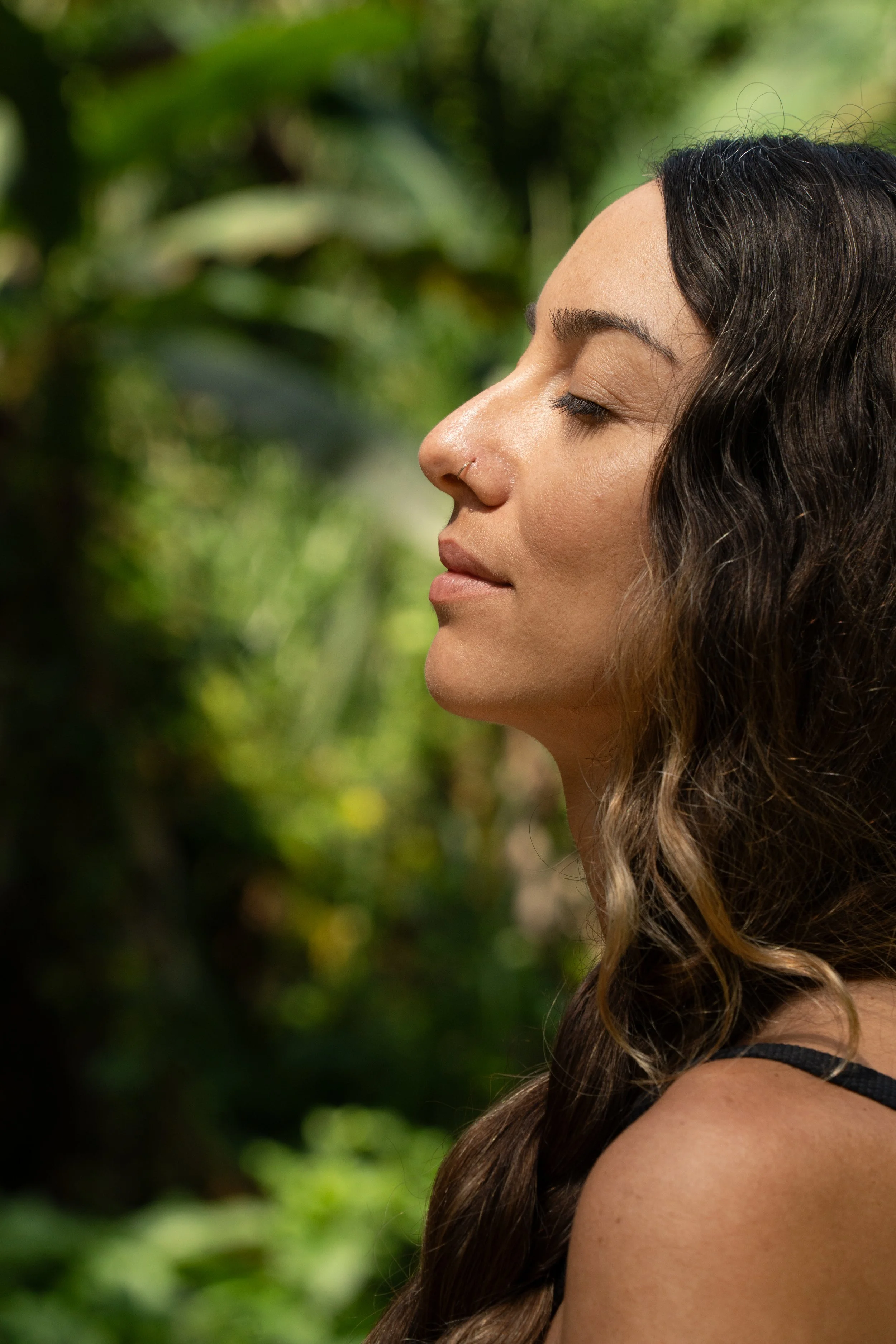 Close-up profile of a woman with closed eyes and long, wavy brown hair, standing outdoors with a blurred green leafy background.