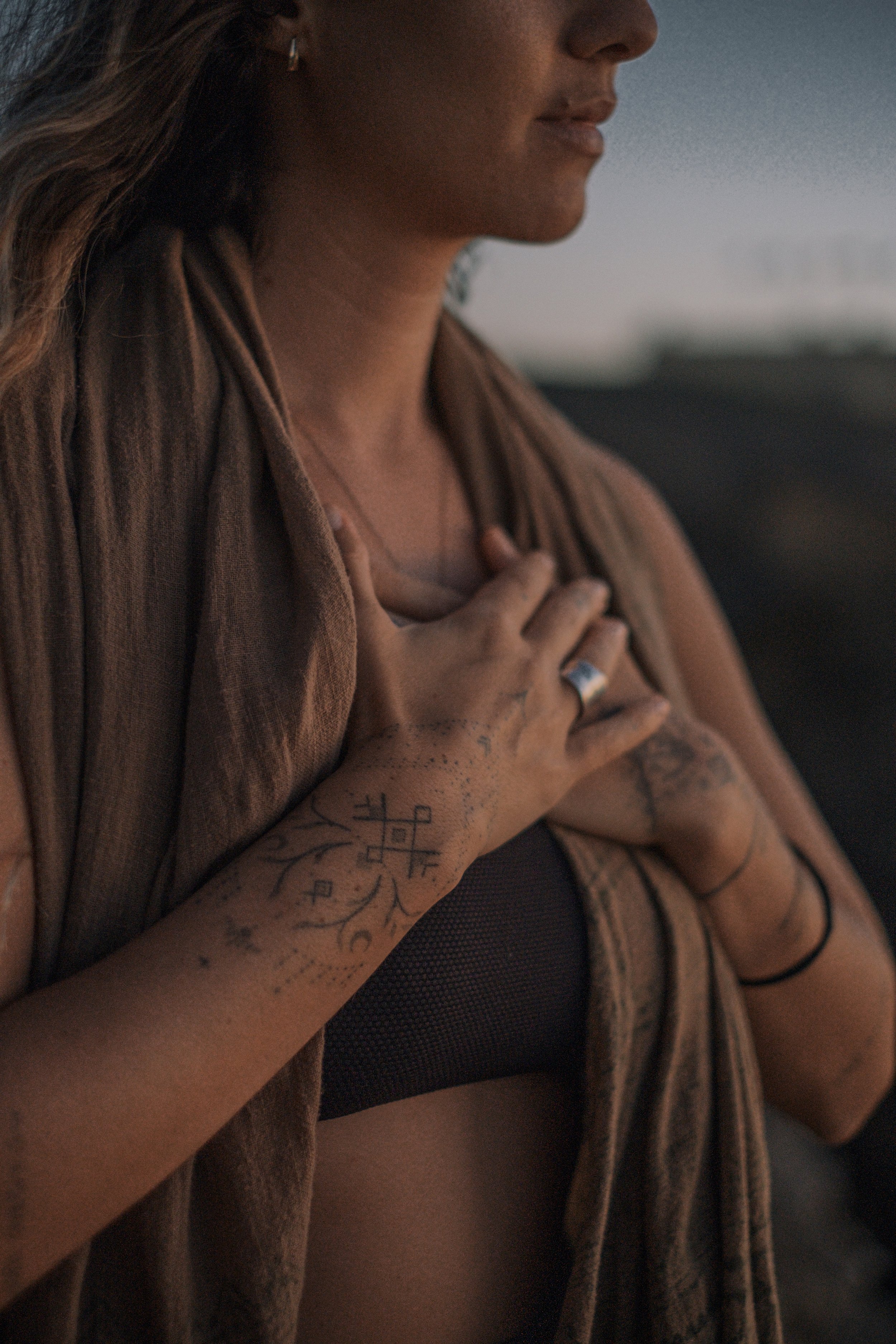 Close-up of a woman with tattoos on her arm, wearing a black top and a brown jacket, with her hand on her chest, standing outdoors at dusk.