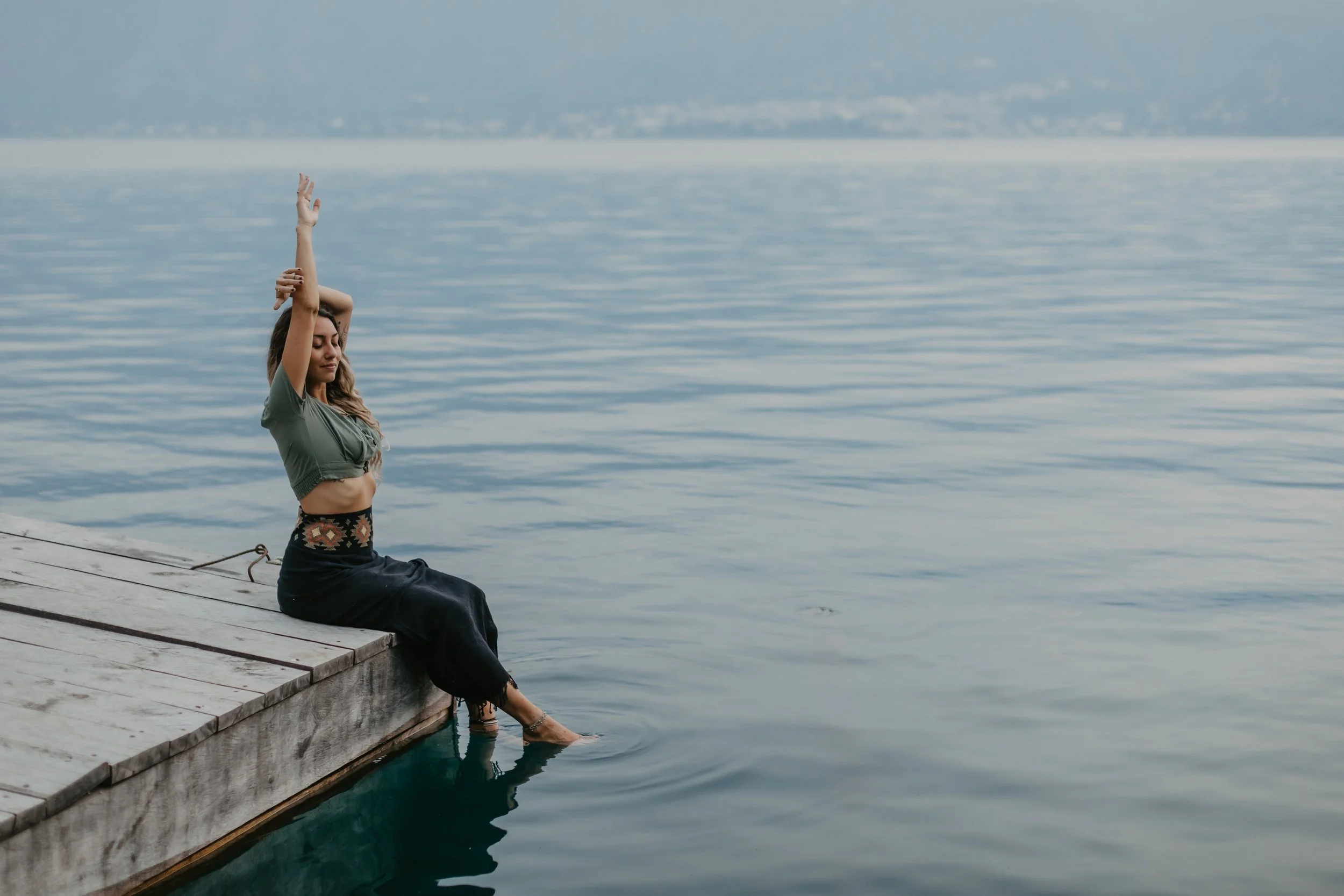 A woman sitting on a wooden dock by the water, with her feet in the water, eyes closed, in a peaceful pose.