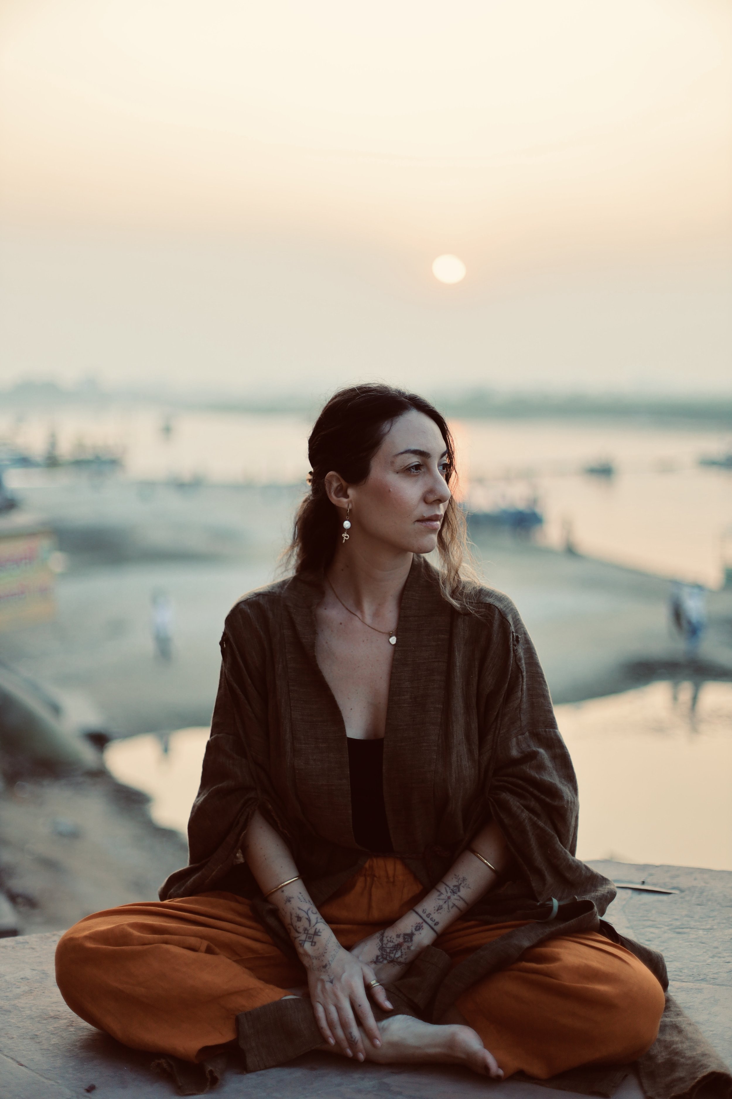 A woman with dark hair, sitting cross-legged on a ledge at the beach during sunset, wearing orange pants, a dark jacket, and displaying tattoos on her arms.