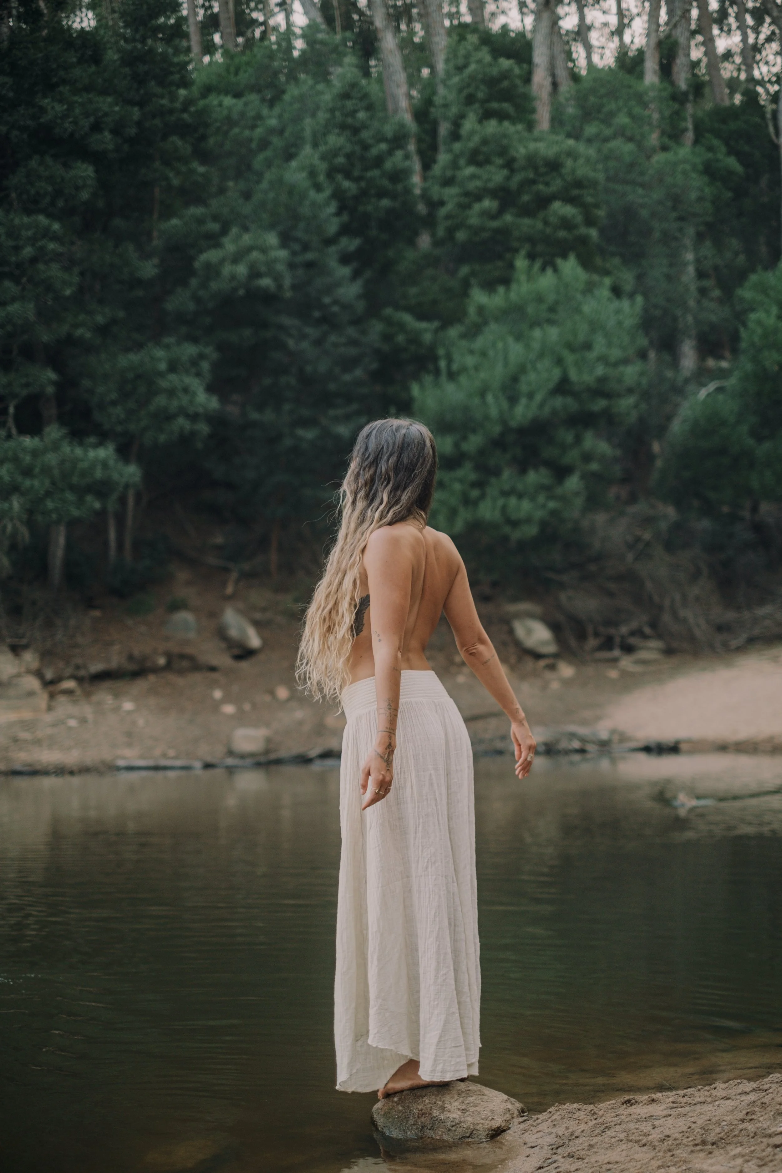 A woman with long wavy hair standing barefoot on a rock in a river, facing away, wearing a white flowing skirt and no top, surrounded by lush green trees.