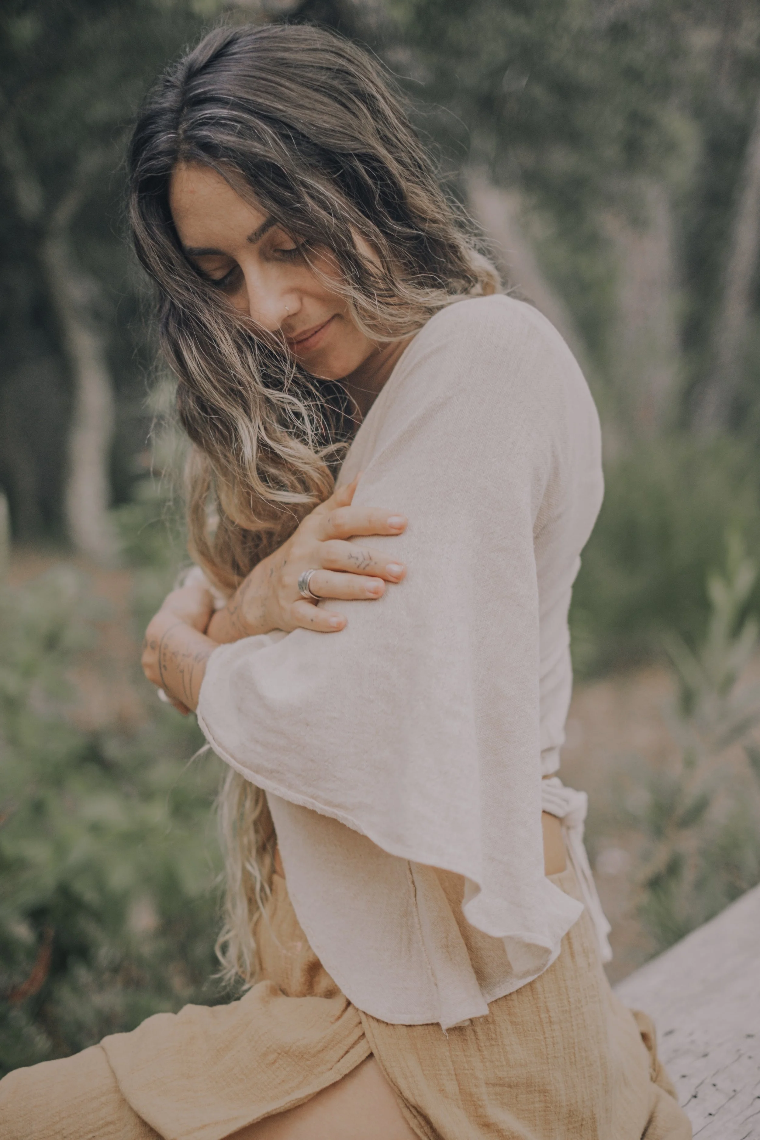 A woman with long hair and tattoos on her arms is embracing herself outdoors, with trees and greenery in the background.