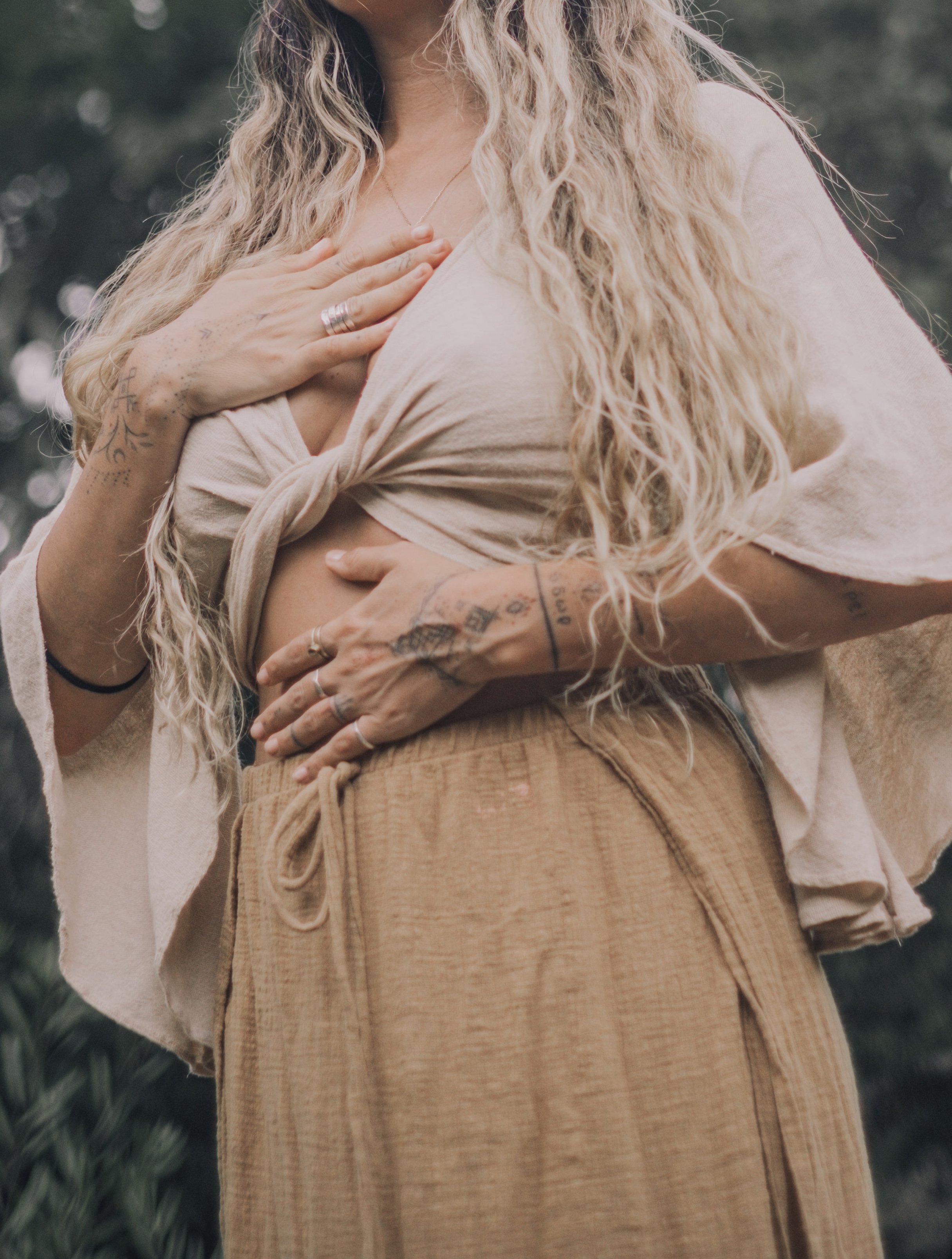 Close-up of a woman with long, curly blonde hair, wearing a white knapped shirt and tan pants, with visible tattoos on her hand and arm, outdoors against a blurred background.