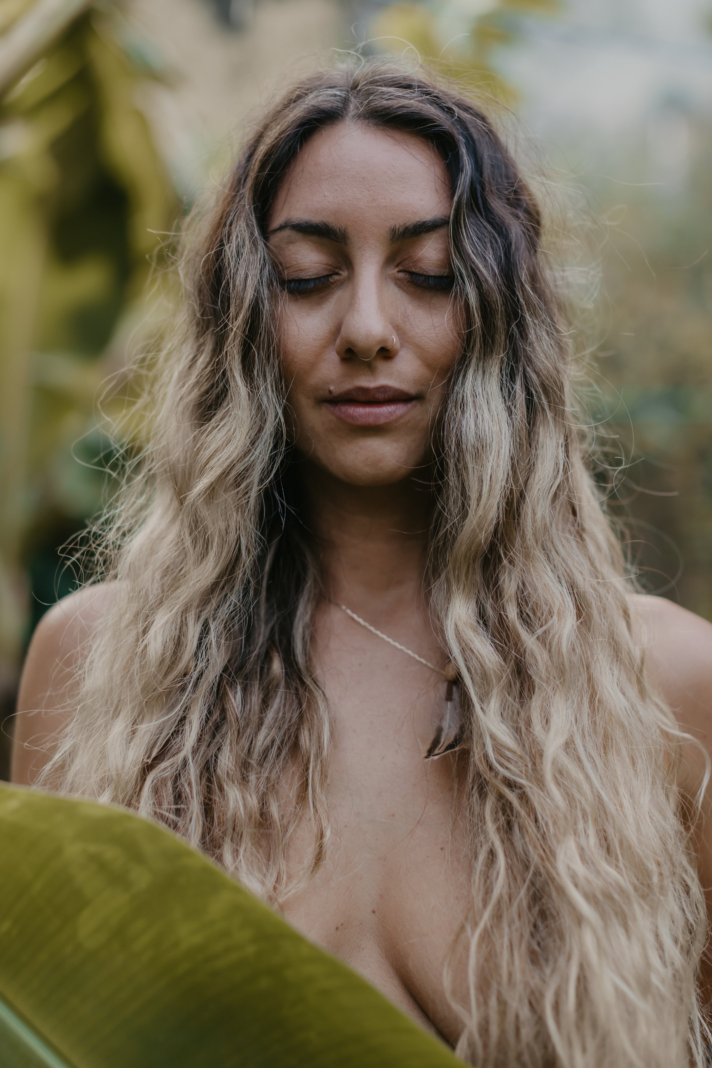 A woman with closed eyes and long, wavy hair standing outdoors near large green leaves.