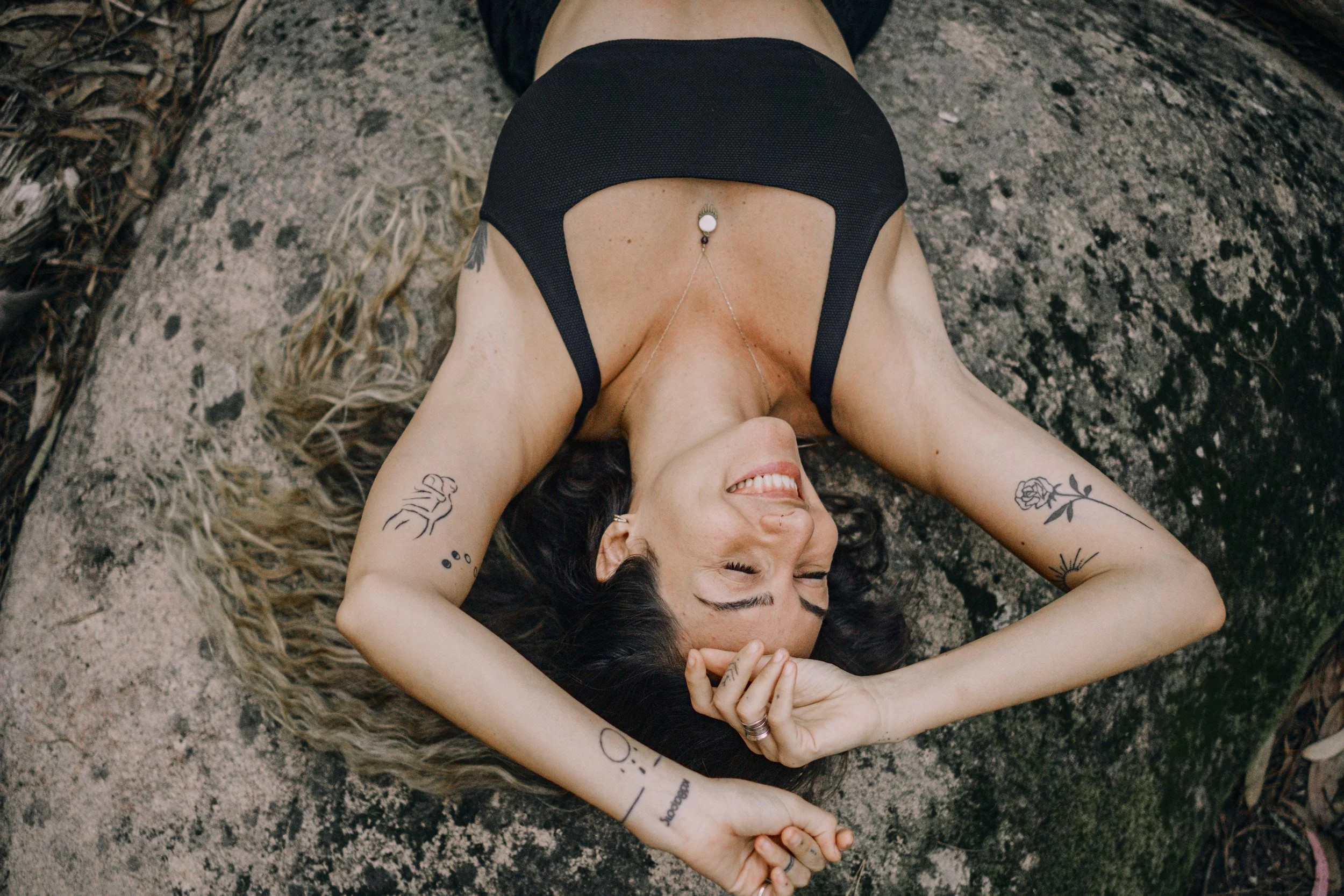 A woman lying on her back on a rocky outdoor surface, smiling with closed eyes, with long dark hair and tattoos on her arms.