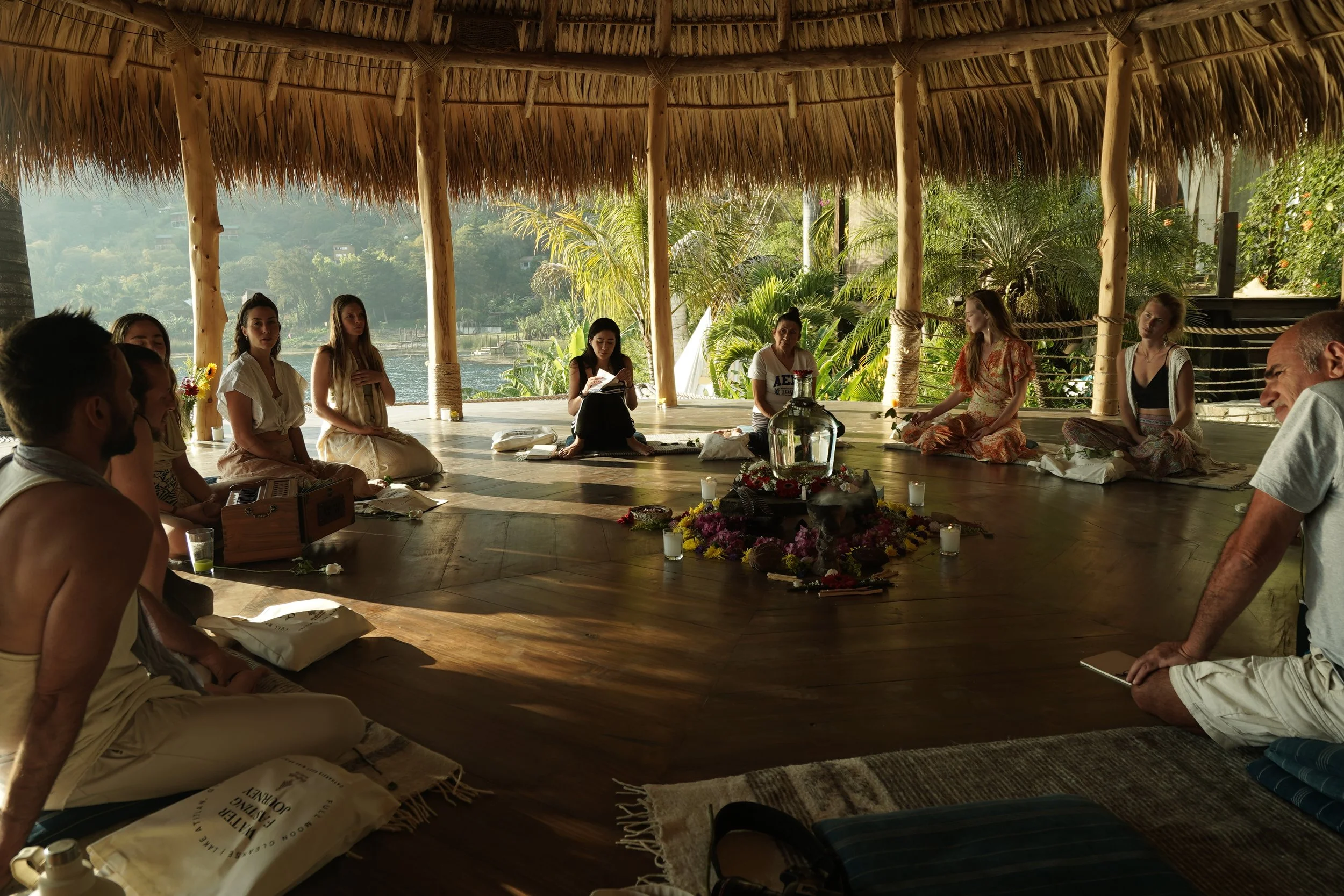 Group of people sitting in a circle under a thatched roof with tropical greenery in the background, participating in a discussion or meditation around a central arrangement of flowers and candles.