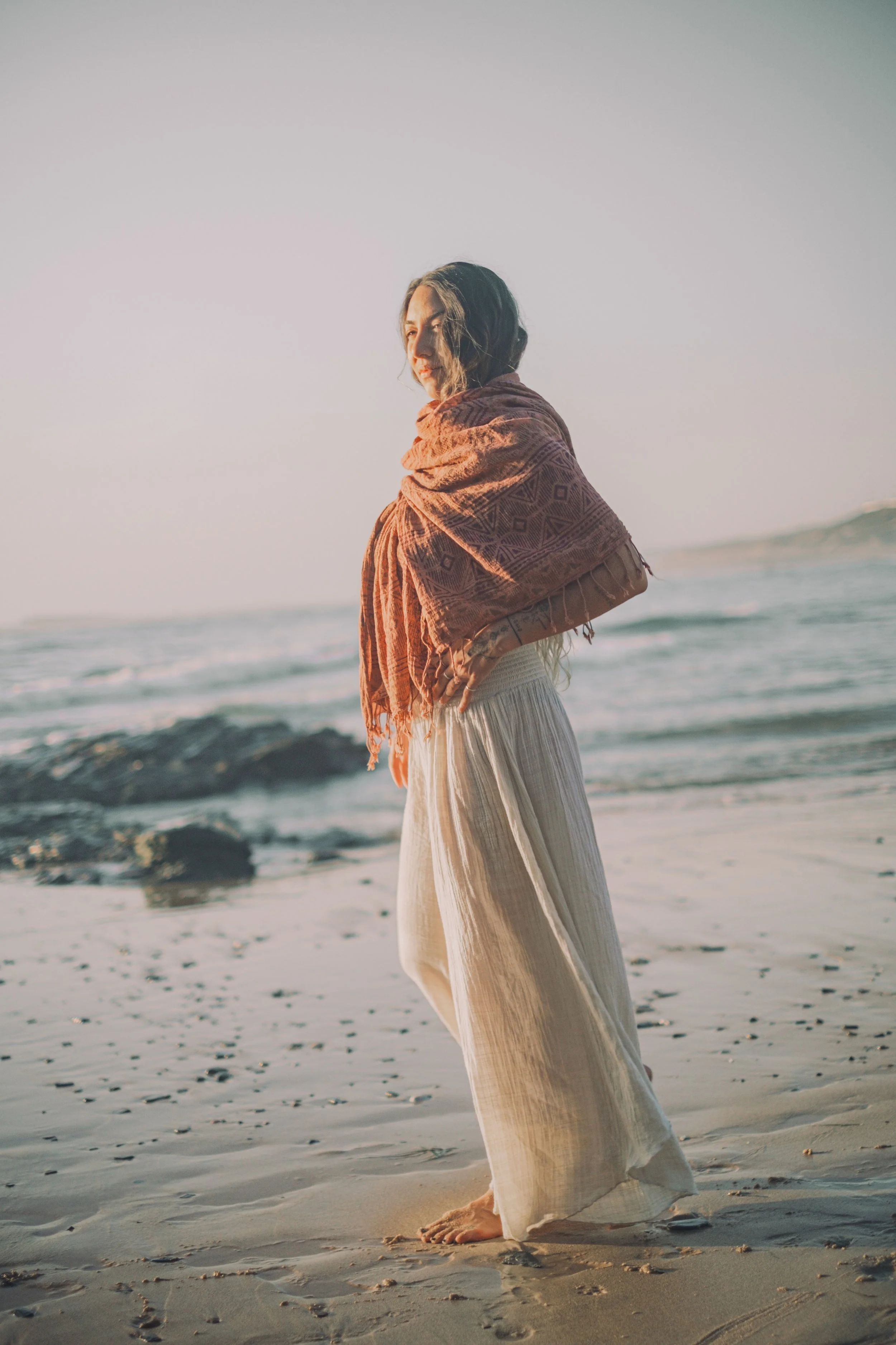 A woman standing barefoot on a sandy beach, wrapped in a pink scarf, gazing pensively at the ocean during sunset.