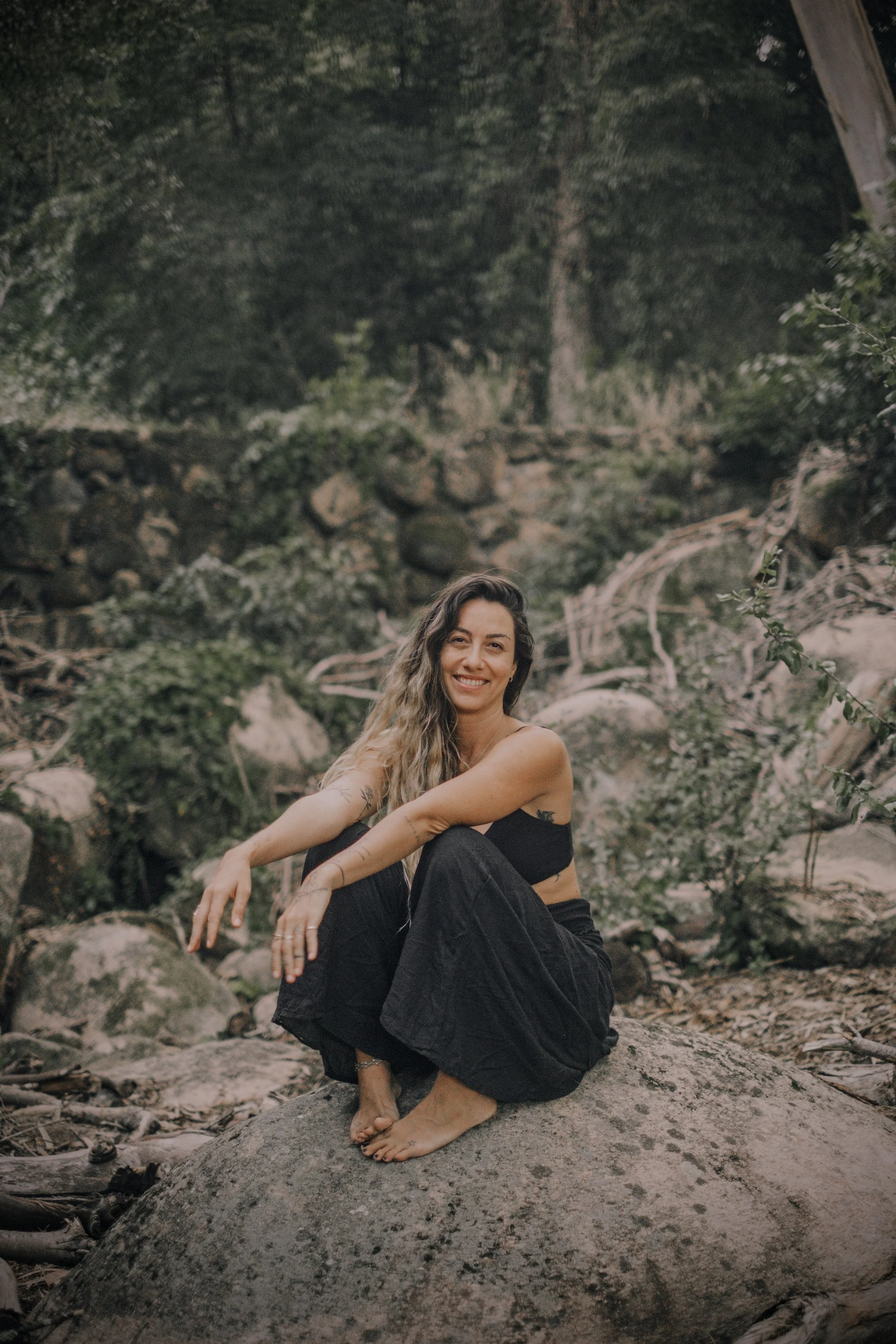 A woman with long wavy hair sitting on a large rock in a natural outdoor setting with trees and rocks, smiling at the camera.