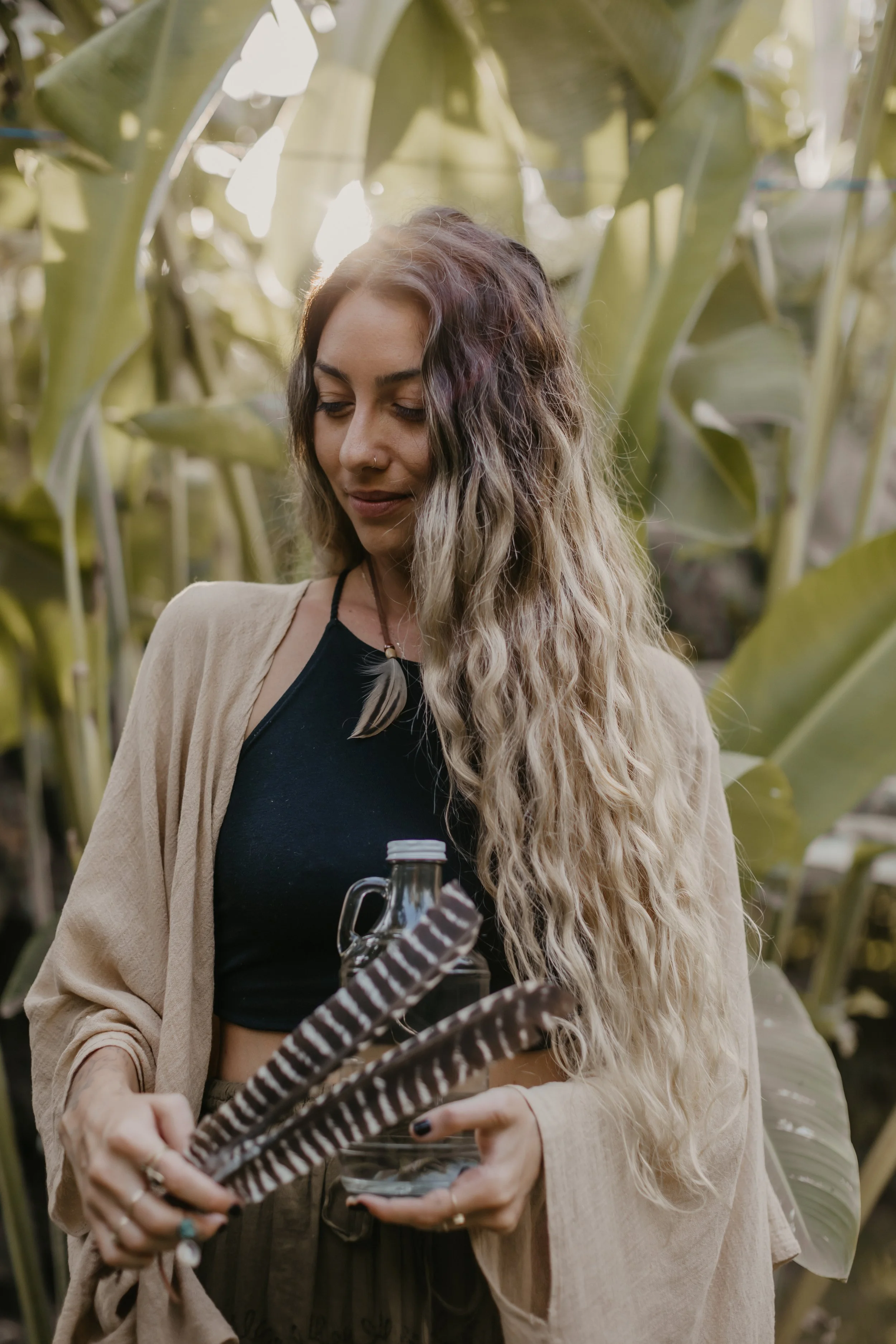 A woman with long wavy hair and a nose piercing holding a glass jar with a black-and-white striped straw, standing among tall green plants in a well-lit outdoor setting.