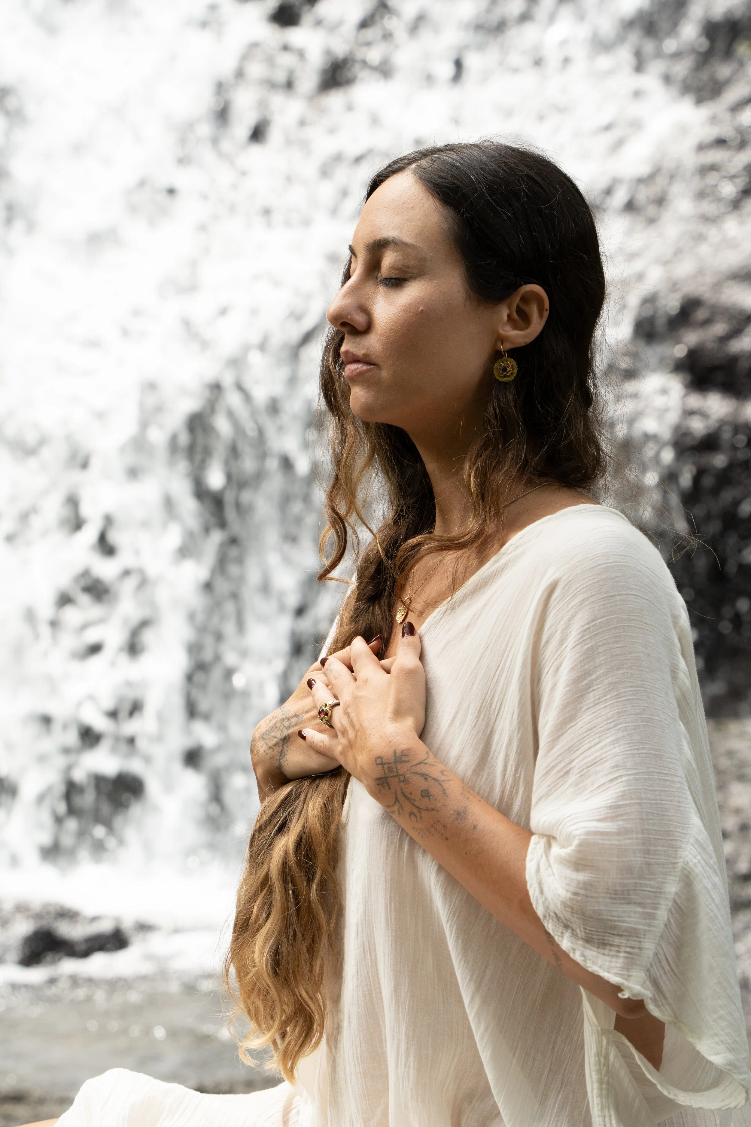 A woman with long wavy hair and tattoos on her hands and arms practices meditation with her eyes closed in front of a waterfall.