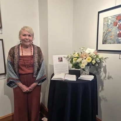 An elderly woman standing next to a table with a flower arrangement and an open book, inside an art gallery or exhibition space.