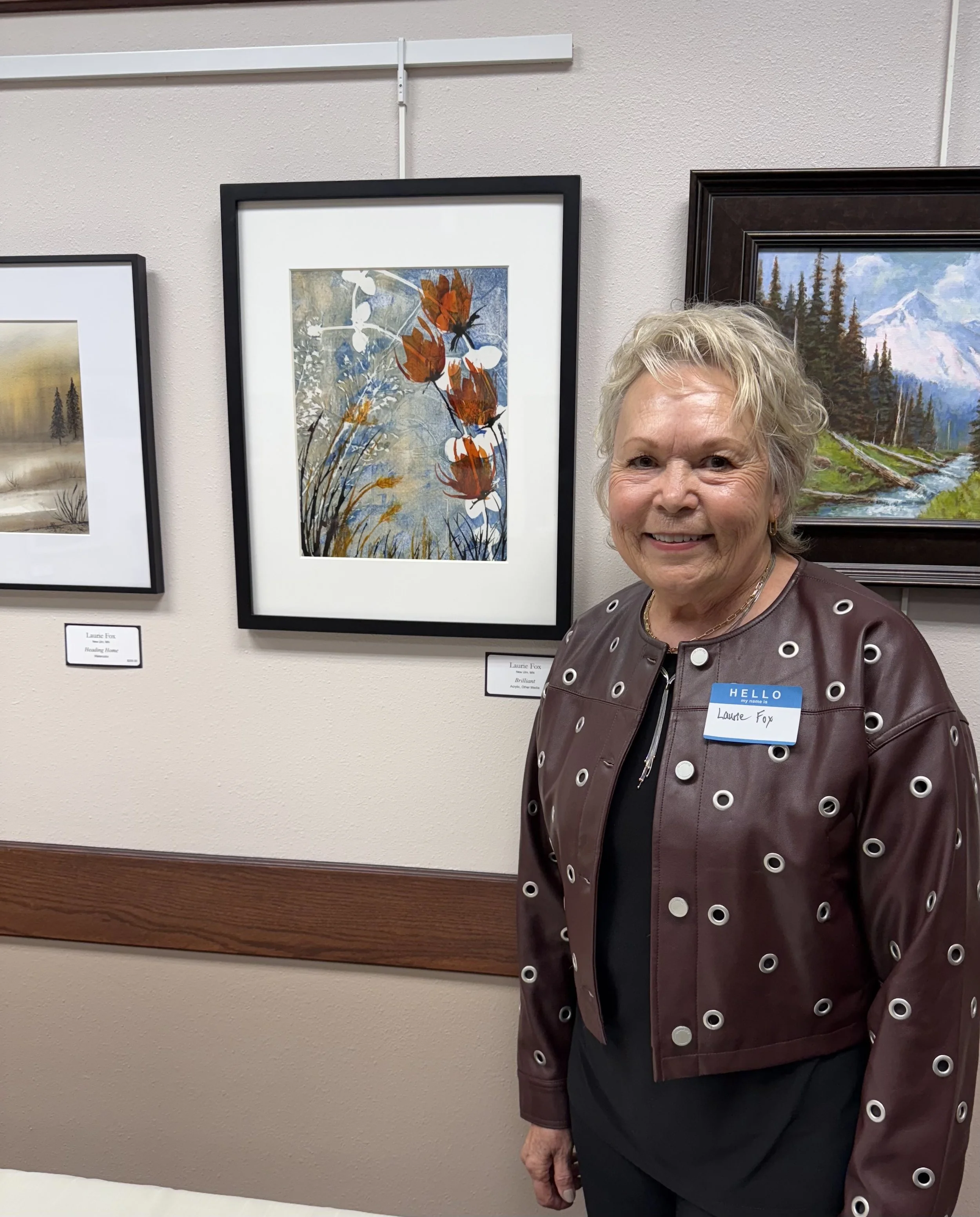 A woman with short gray hair smiling, wearing a dark top and a maroon jacket with large silver grommets, standing in front of framed artwork on a beige wall at an art gallery or exhibition.