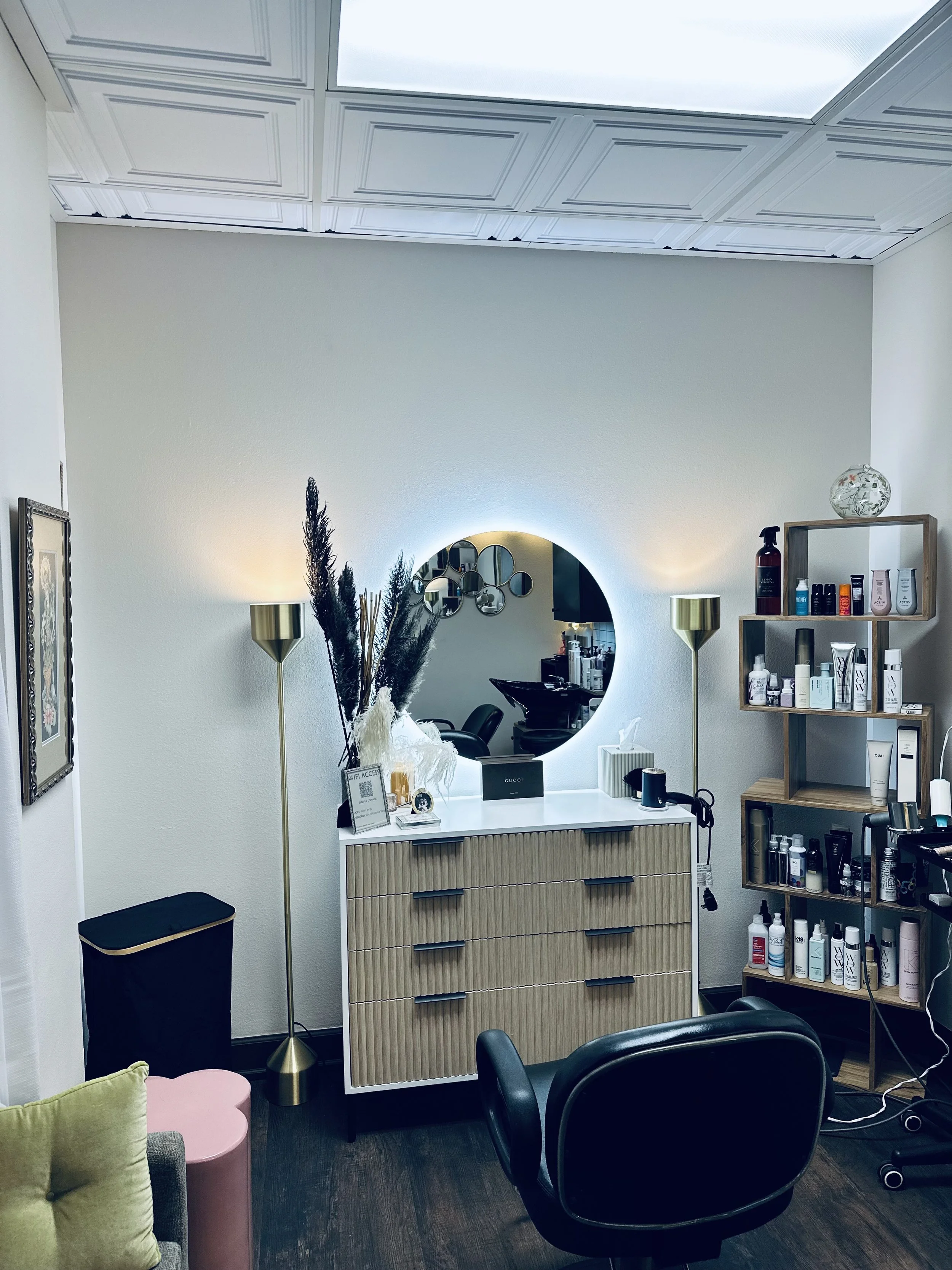 Interior of a hair salon or beauty studio with a white reception desk, round mirror, black chairs, wooden shelving with beauty products, and decorative items like lamps and a vase.