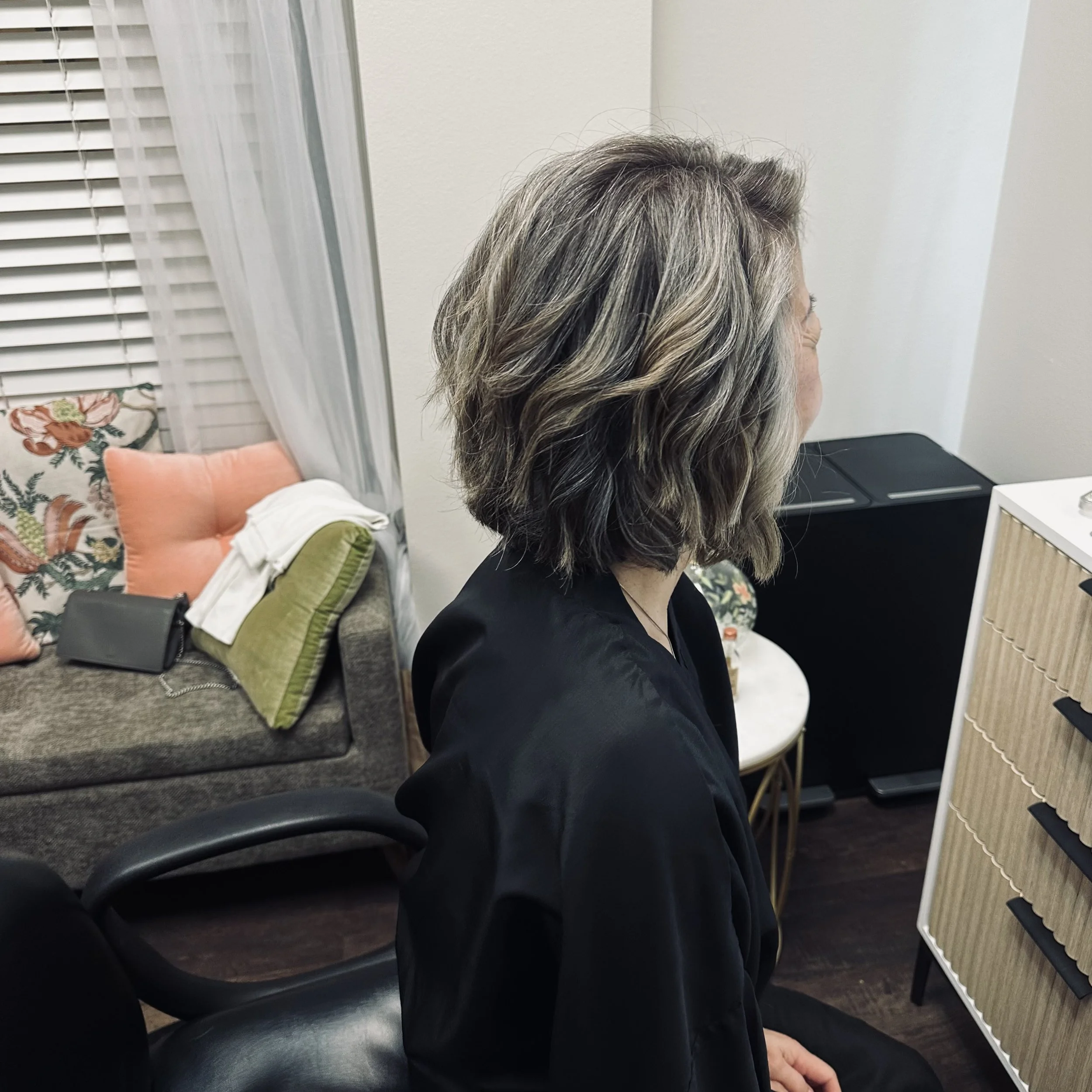 Side view of a woman with gray hair sitting in a waiting room, with a black chair, orange and green pillows, and window blinds in the background.