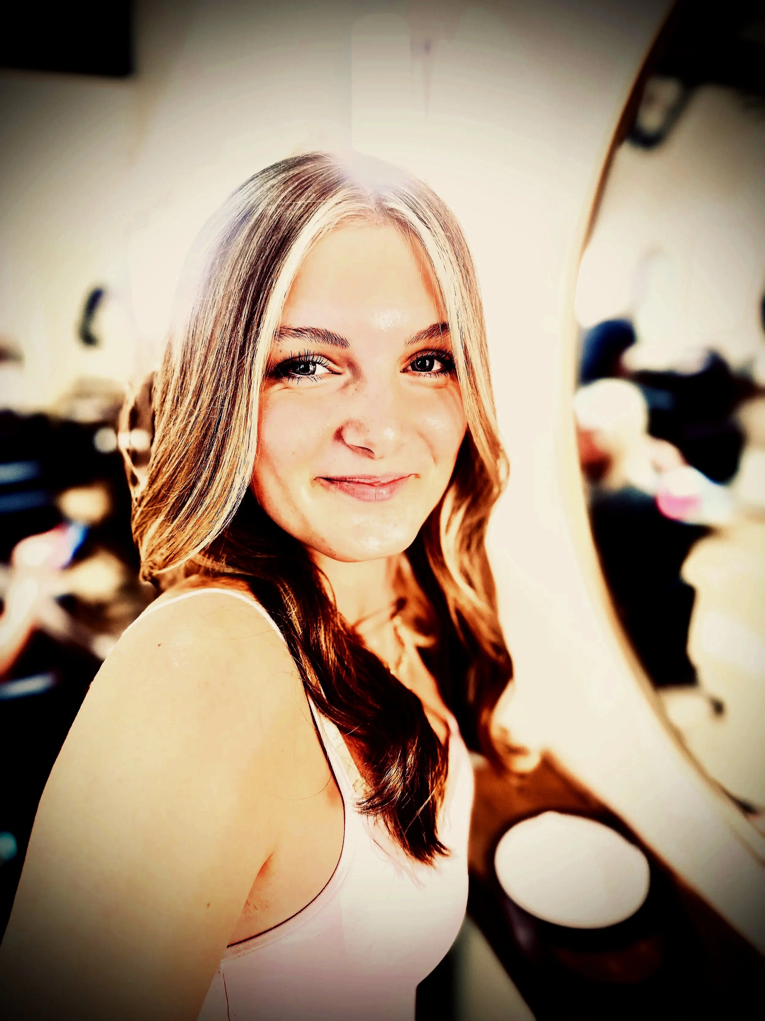 Close-up of a young woman with long, light brown hair and light skin, smiling at the camera, with a blurred background showing a person and hanging lights.