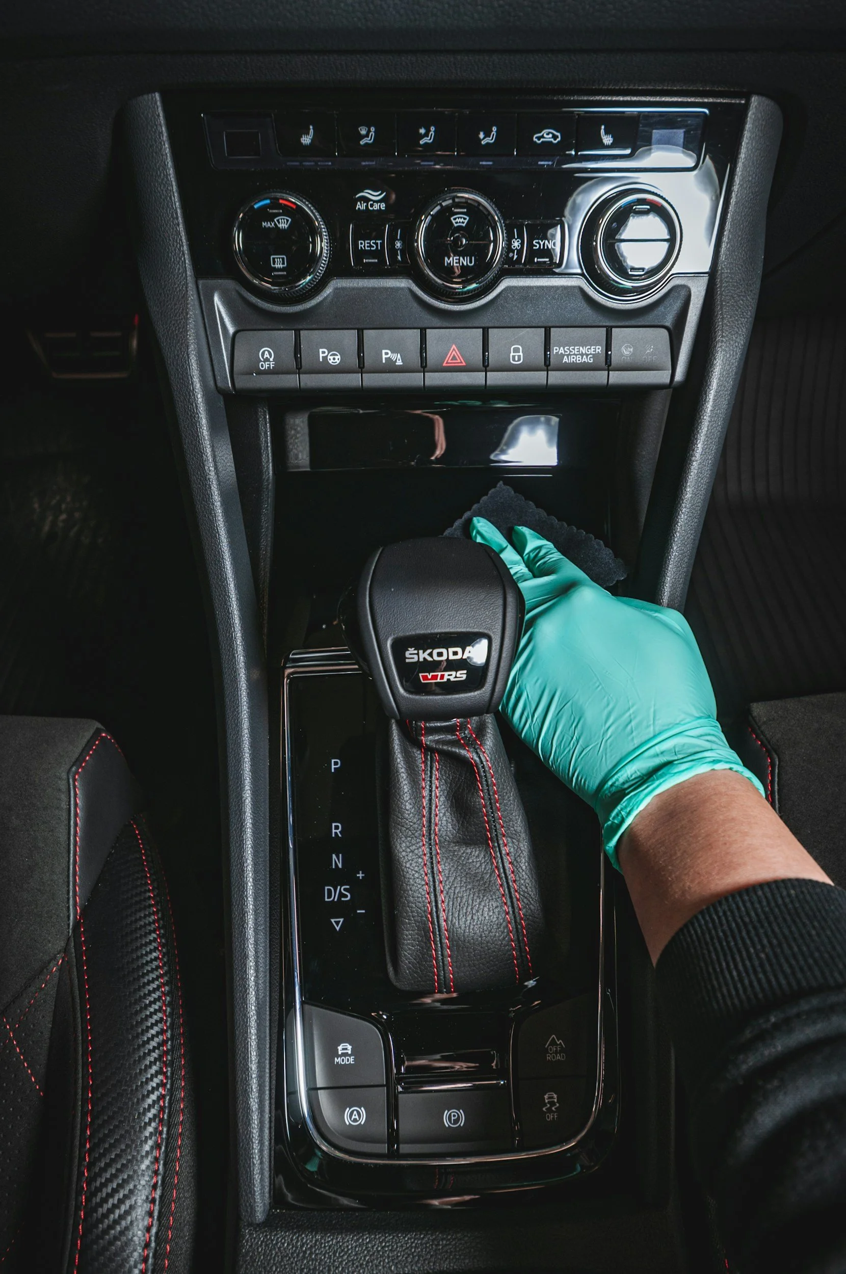 Innenraum eines Autos mit Hand in einer grünen Gummihandschuh beim Hochschalten des Schalthebels, der Markenname Škoda auf dem Schalthebel, Automatikgetriebe, Steuerknöpfe für Klimatisierung, Warnblinkanlage, Parkbremse und andere Funktionen.
